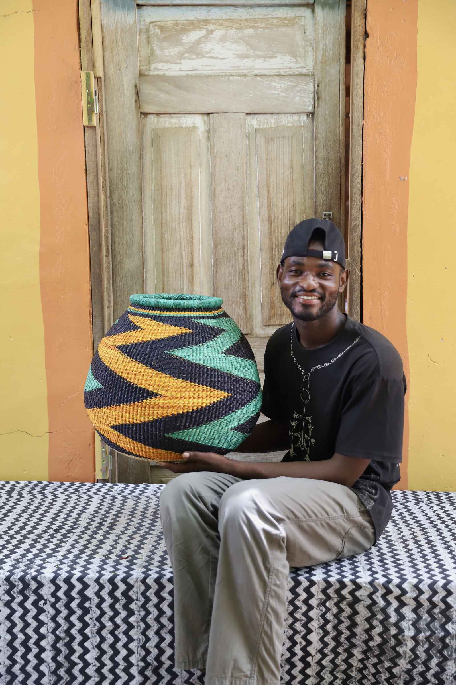 Handwoven African art basket with bold diagonal zigzag pattern in teal, black and amber, held by a man in Bolgatanga.
