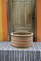 Handwoven Bolga storage basket in natural straw with orange and black horizontal stripes and twin handles.