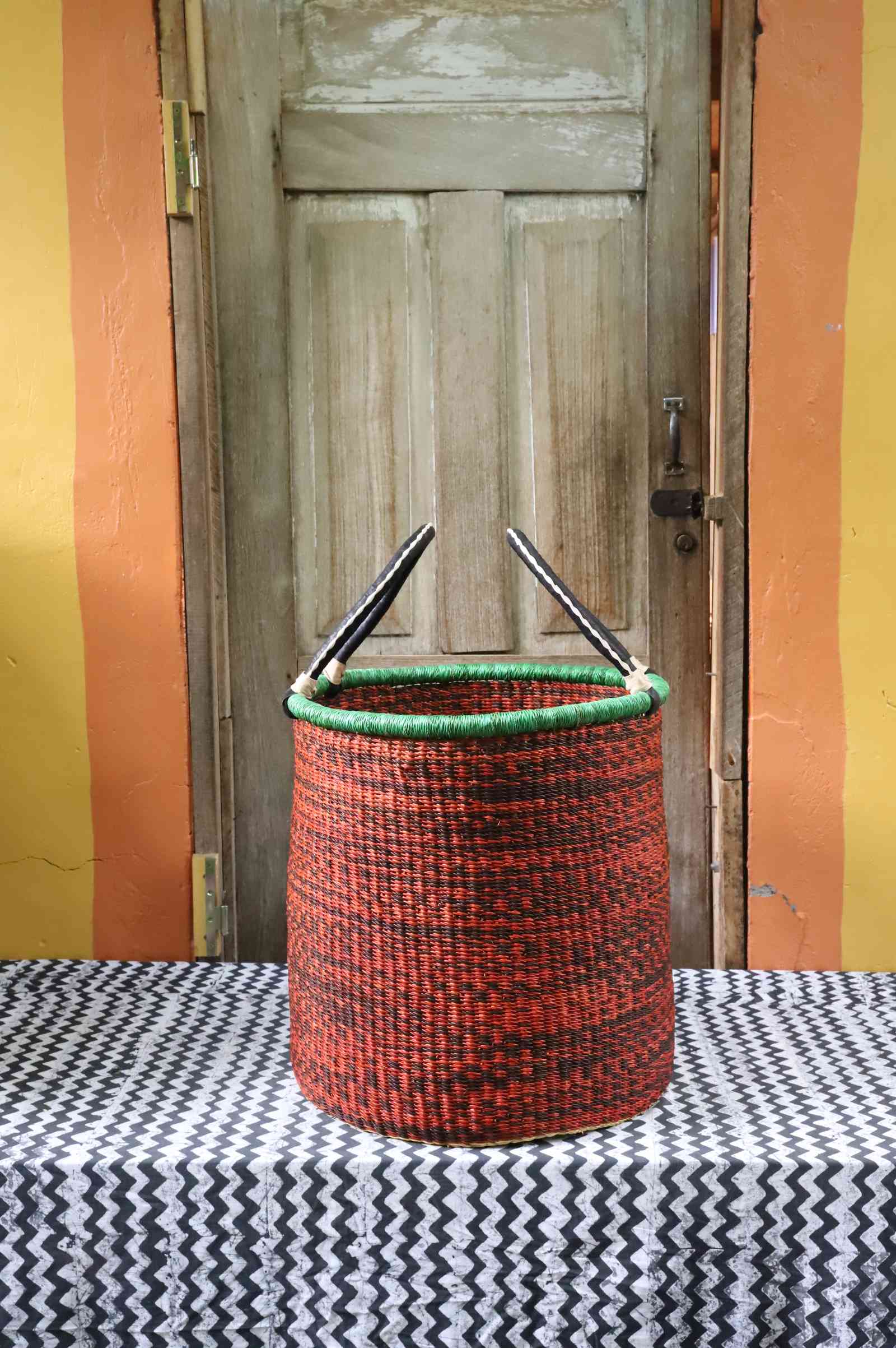 Tall open Bolga laundry basket in speckled rust-red weave with a green rim edge and leather-topped handles.