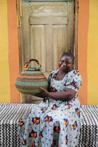 Handwoven Bolga pot basket in multicoloured bands of green, teal and amber, held by a woman seated in Bolgatanga.