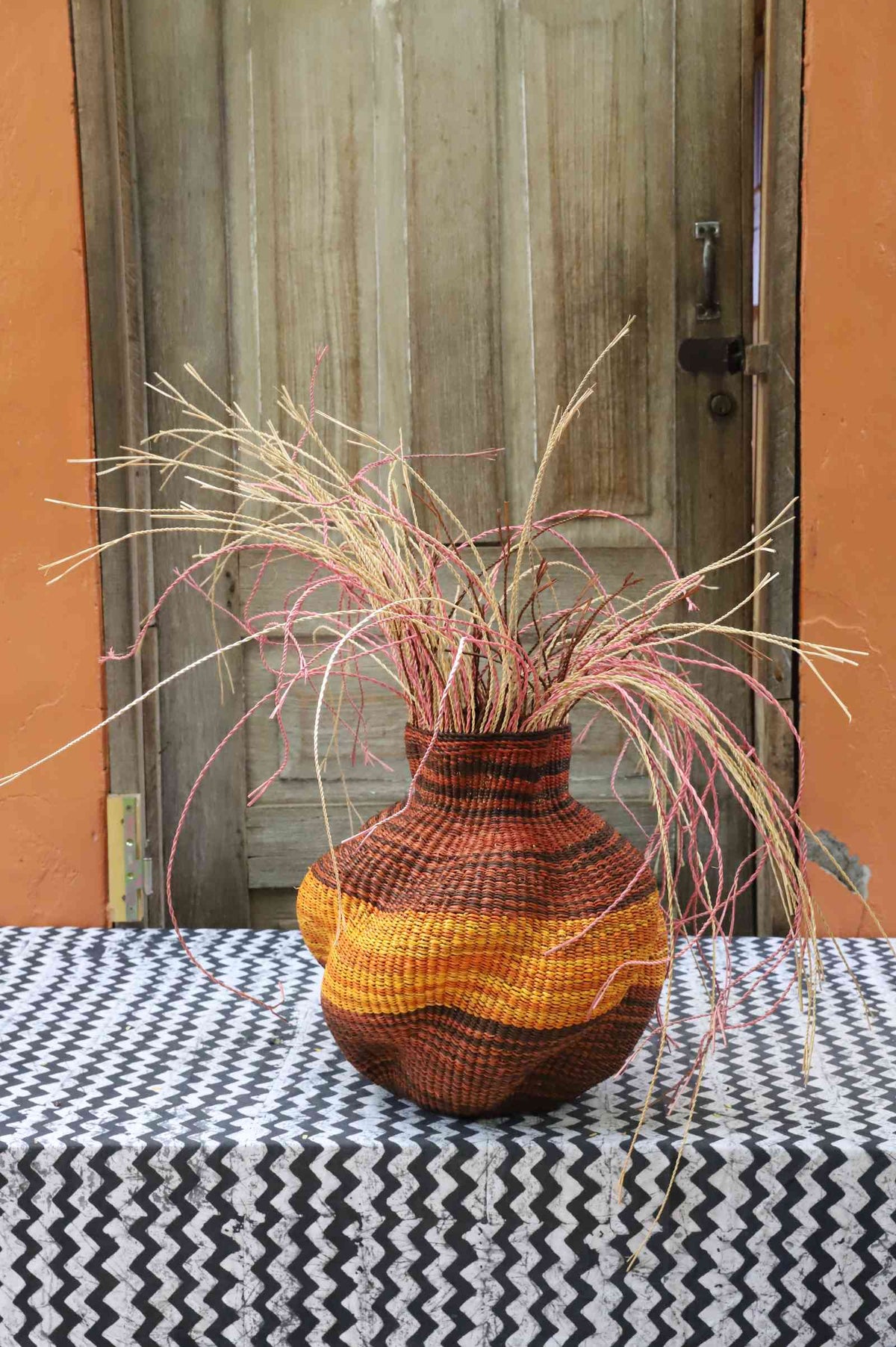 Handwoven Bolga Baobab-inspired basket with dark brown base, orange banded shoulder and pink grass sprigs.