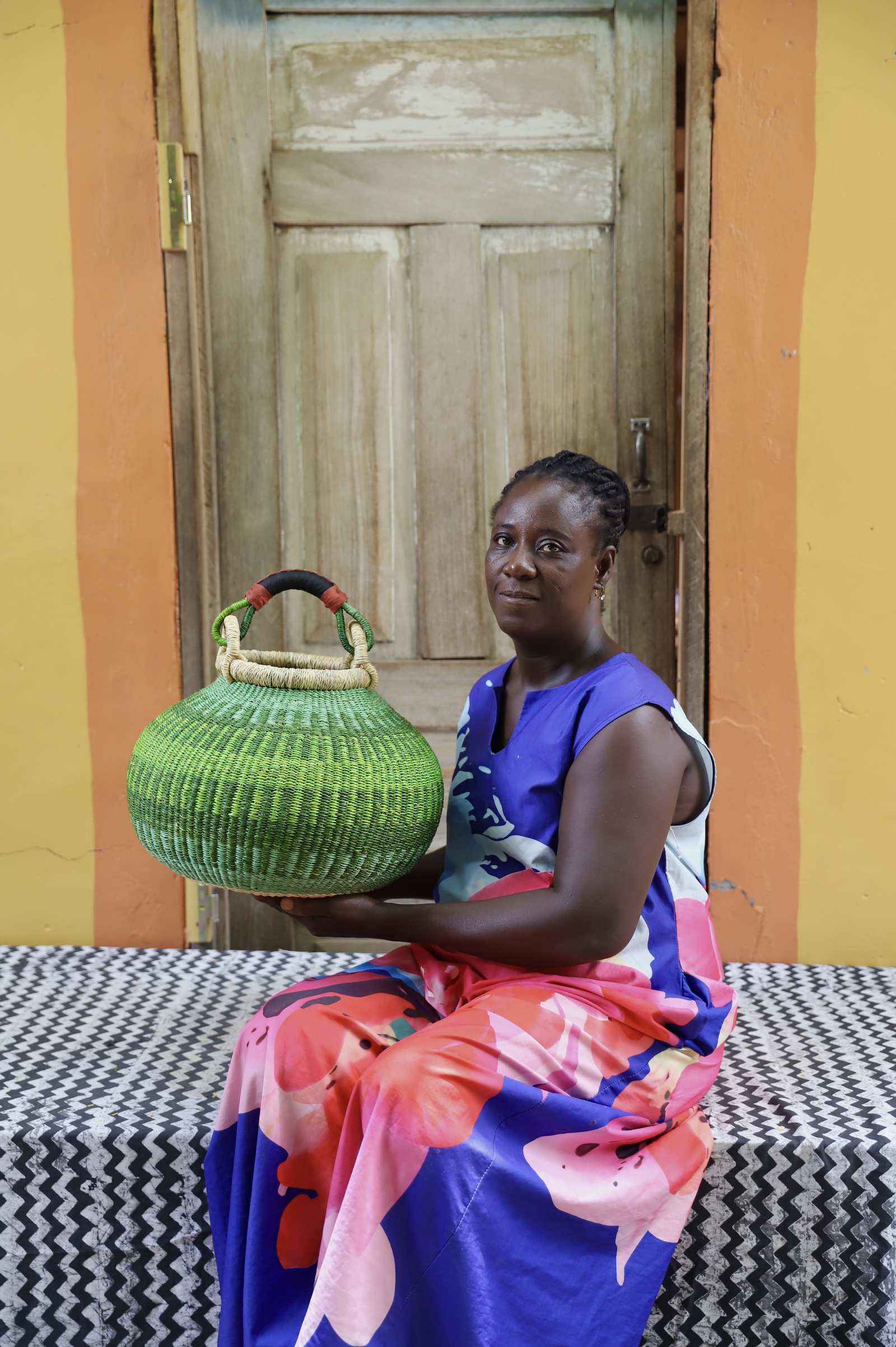 Large handwoven Bolga pot basket in lime green elephant grass with leather top handle, held by a woman seated in Bolgatanga.