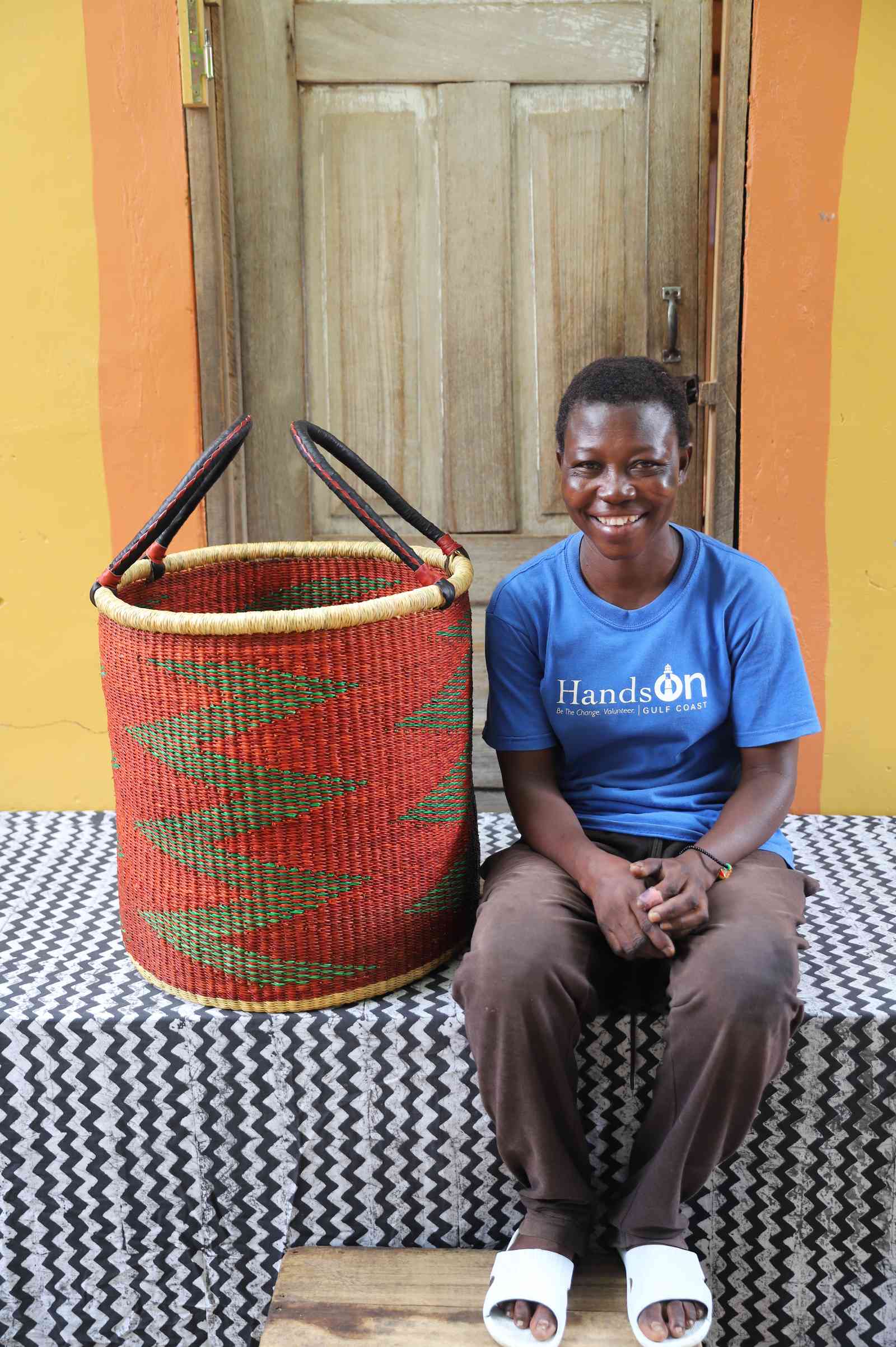 Large open laundry basket in red and green zigzag weave, shown beside a seated person for scale.