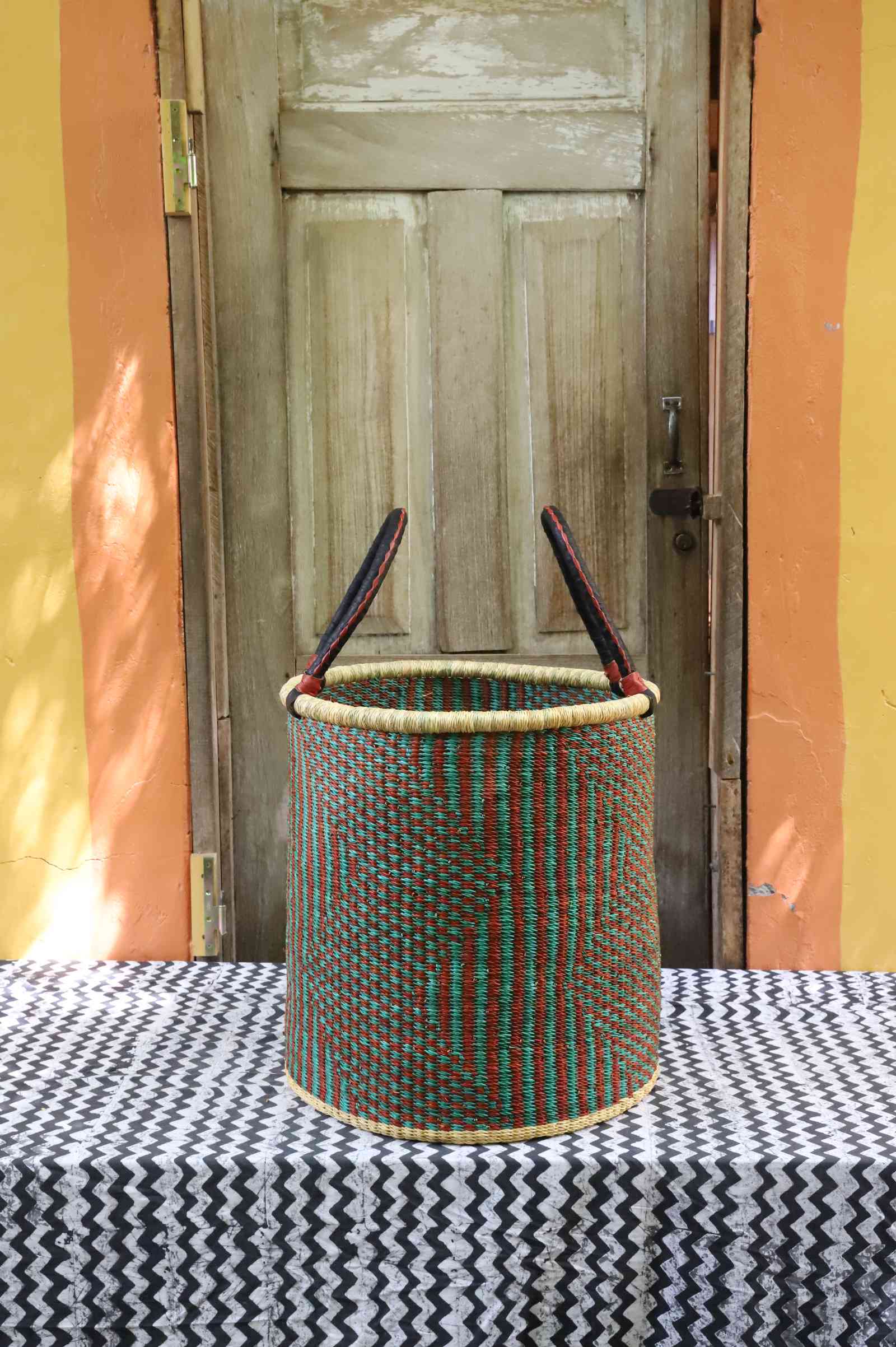 Tall open Bolga laundry basket in a green and rust striped weave with leather-topped handles, on a tablecloth.