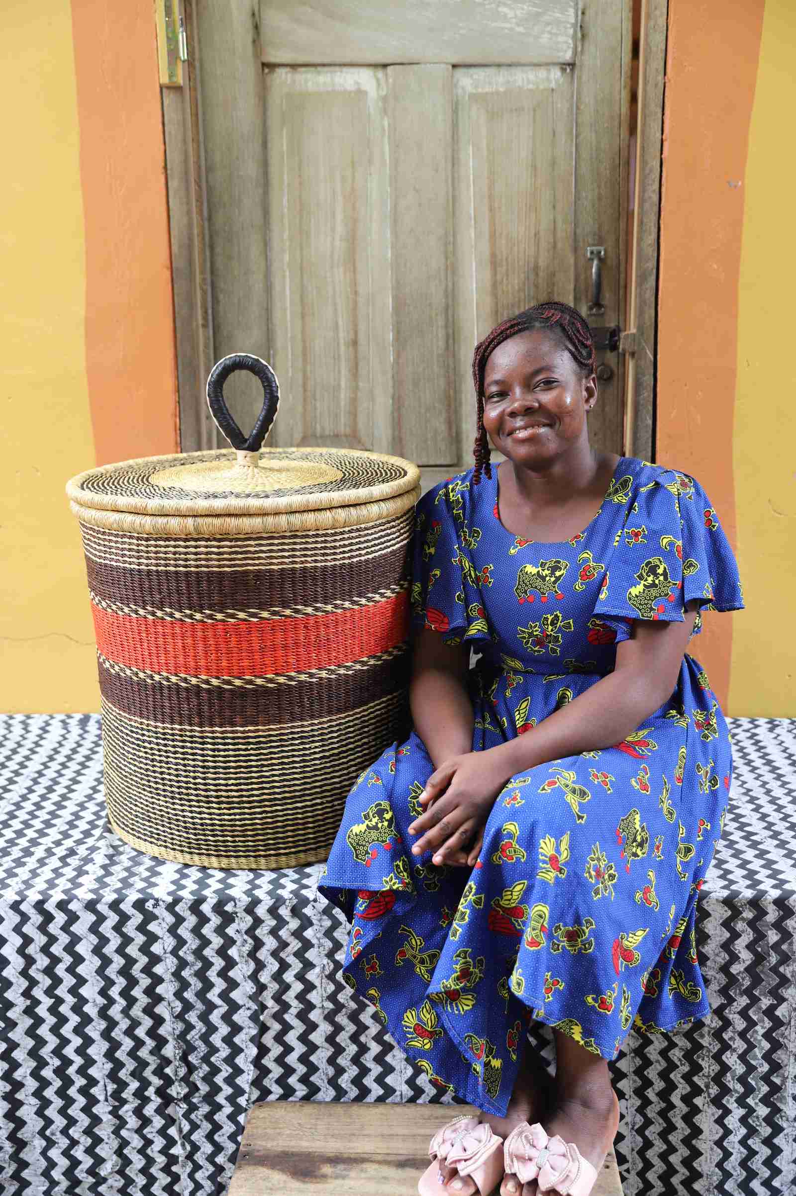 Large coiled-rim lidded laundry basket in natural straw with a red and brown band, shown beside a seated person for scale.