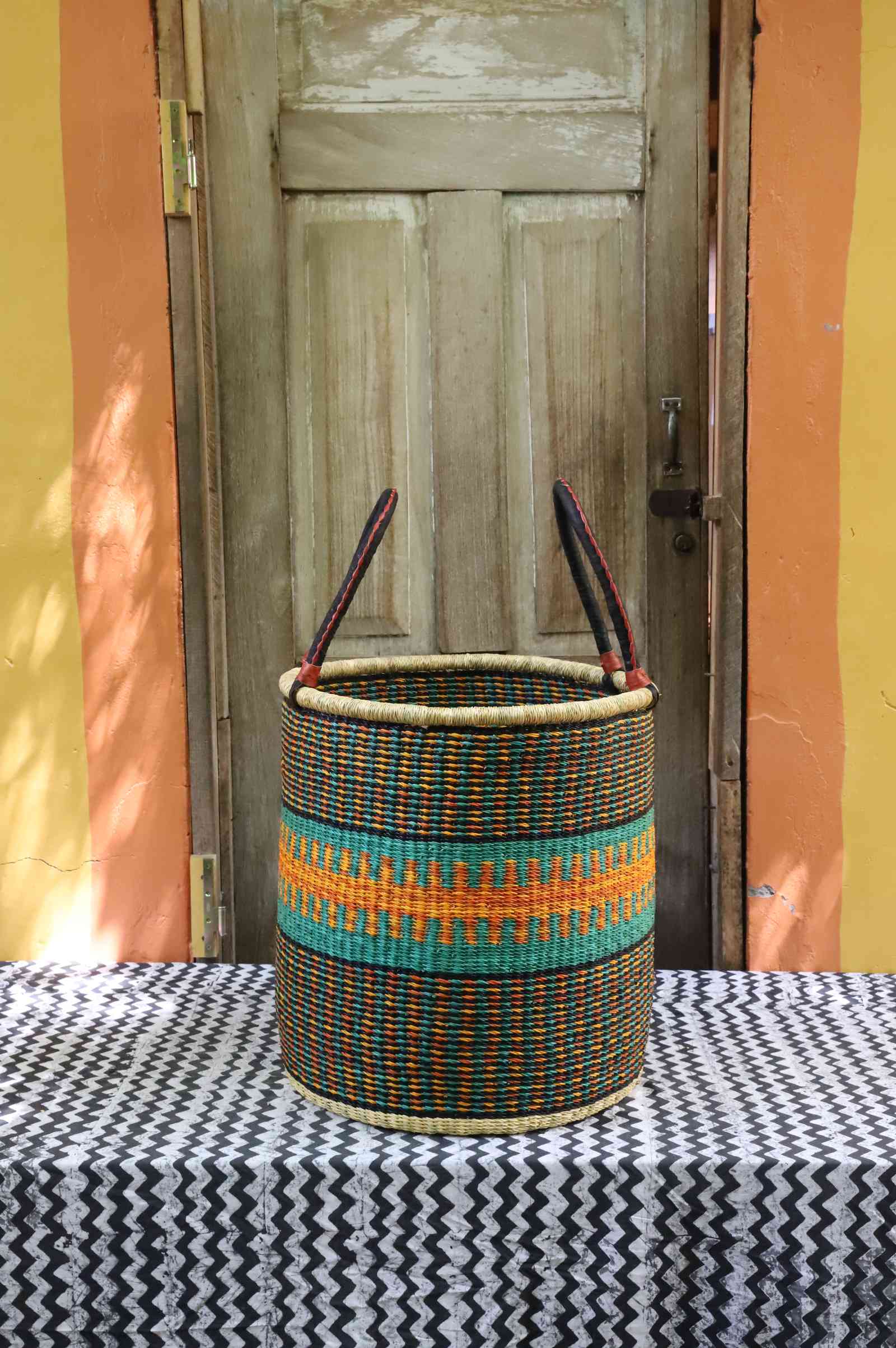 Tall open Bolga laundry basket in dark weave with a teal and orange patterned band and tall leather-topped handles.
