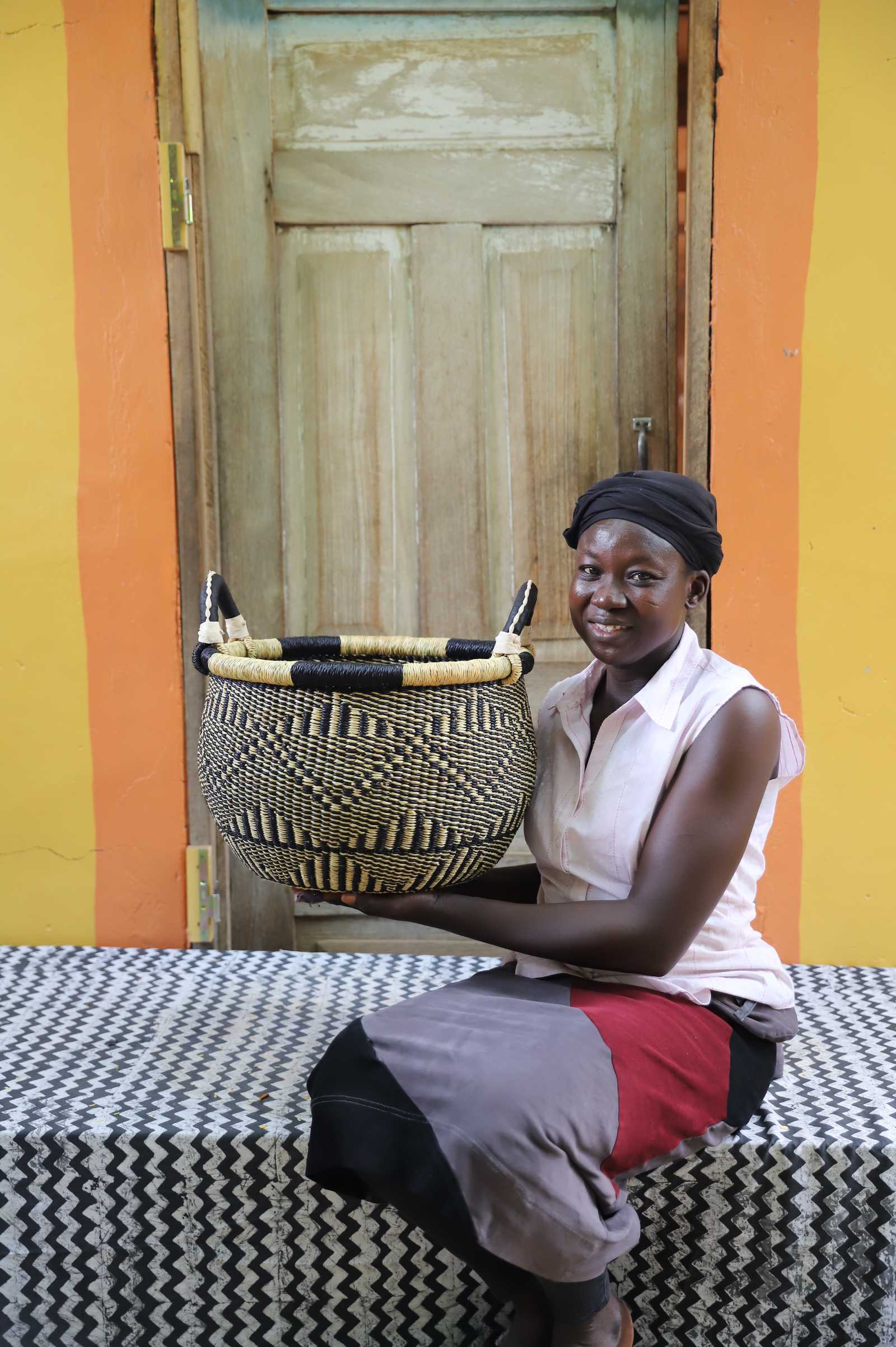 Handwoven Bolga storage basket in natural straw with black diamond pattern, held by a woman against a yellow wall.