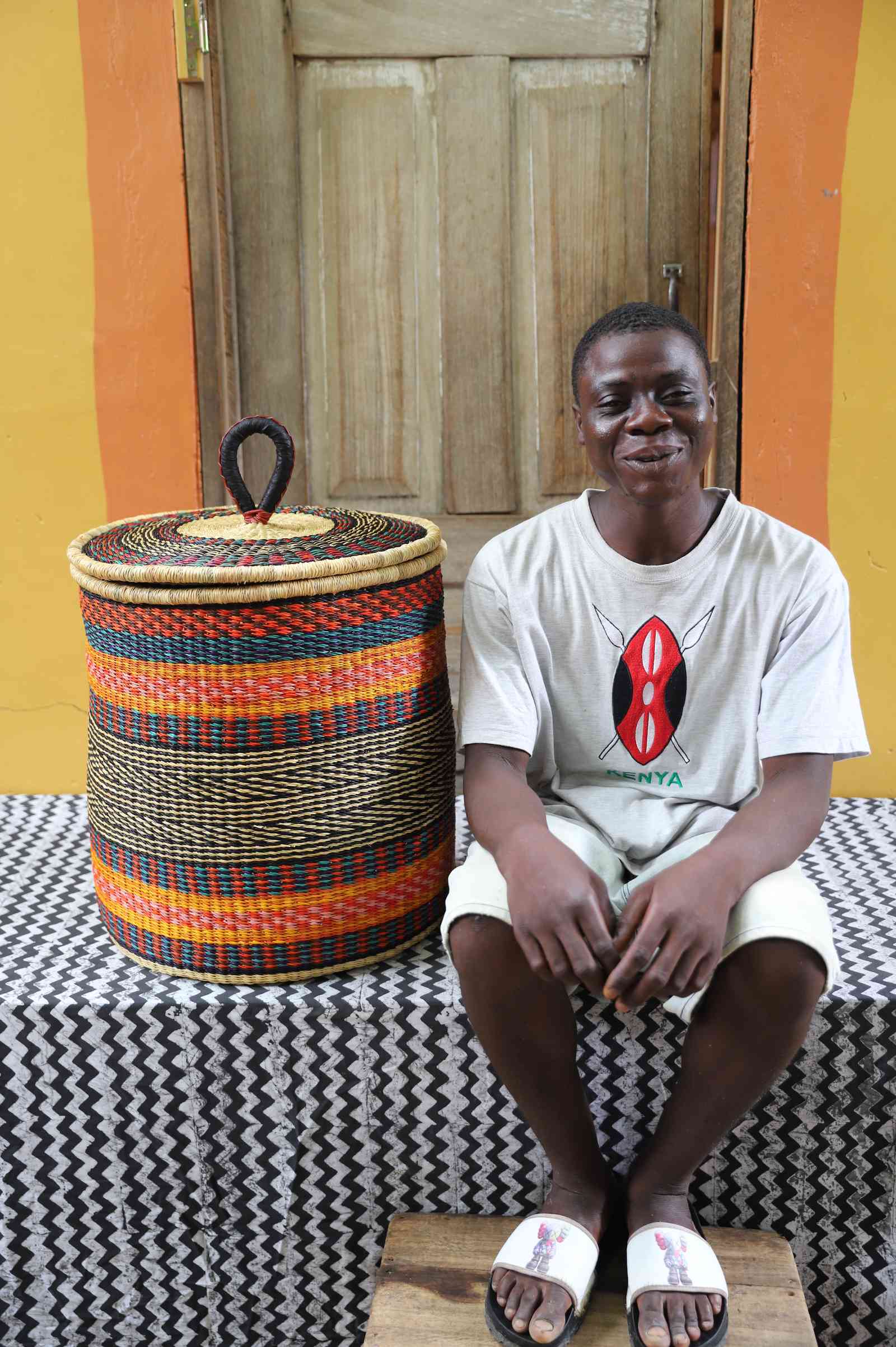 Coiled-rim lidded laundry basket in orange, red and green striped weave, shown beside a seated person for scale.