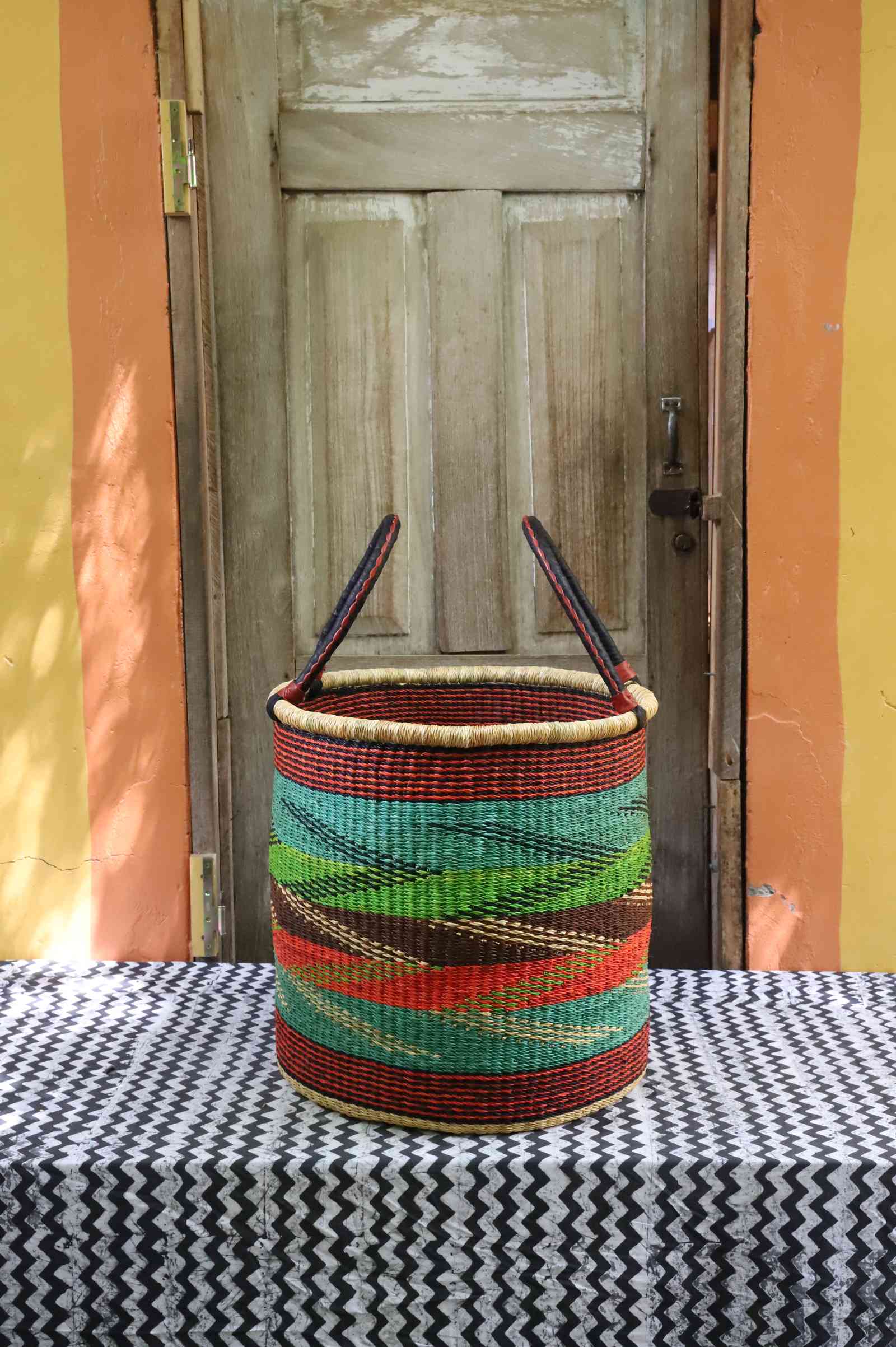 Tall open Bolga laundry basket with a multicolour diagonal striped weave in teal, green and rust, on a tablecloth.