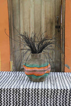 Handwoven Bolga Baobab-inspired basket in natural straw with orange and teal wave stripes and dark grass sprigs.