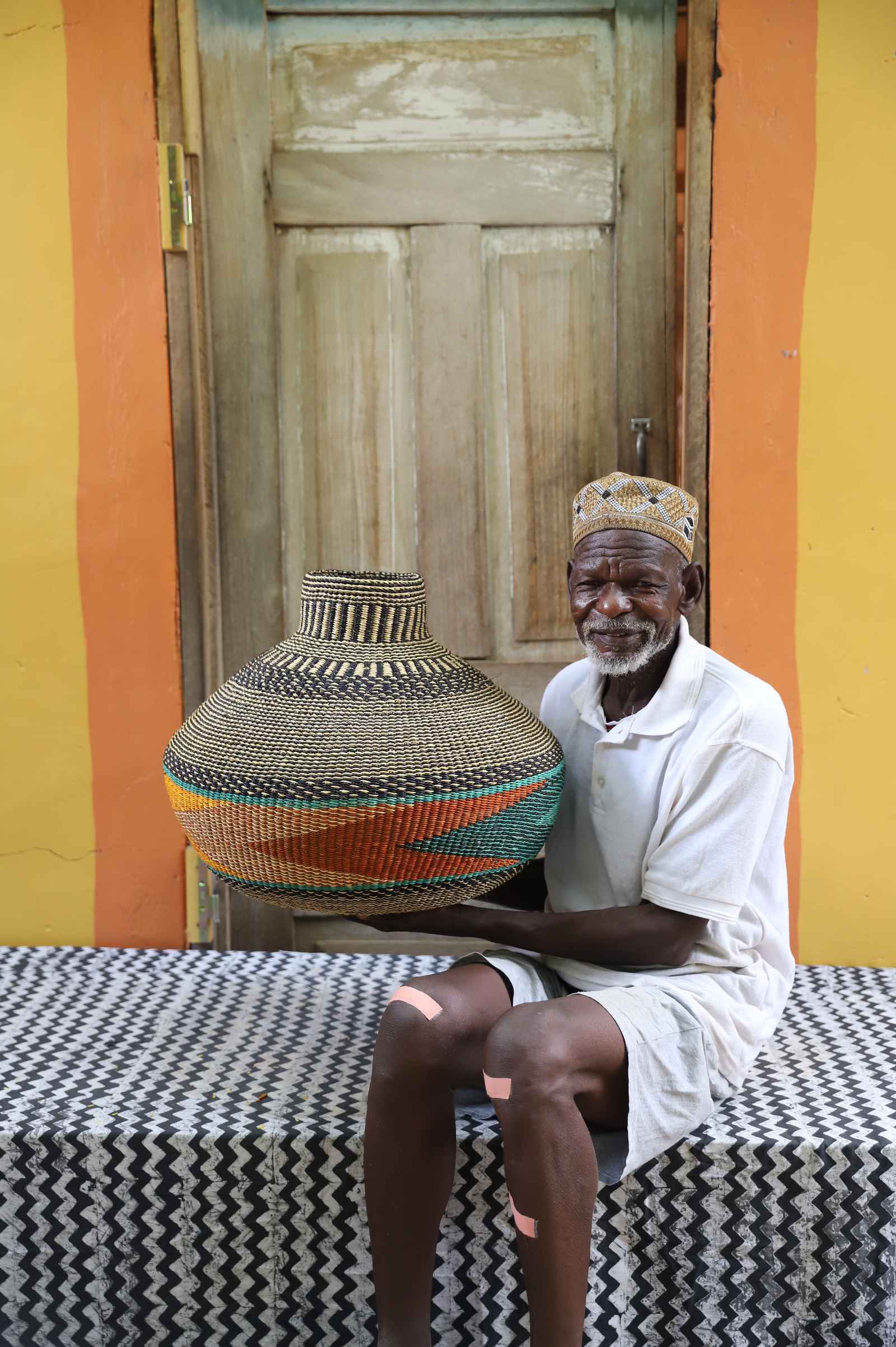 Handwoven African art basket in vase form with terracotta and teal bands, held by a man seated in Bolgatanga.