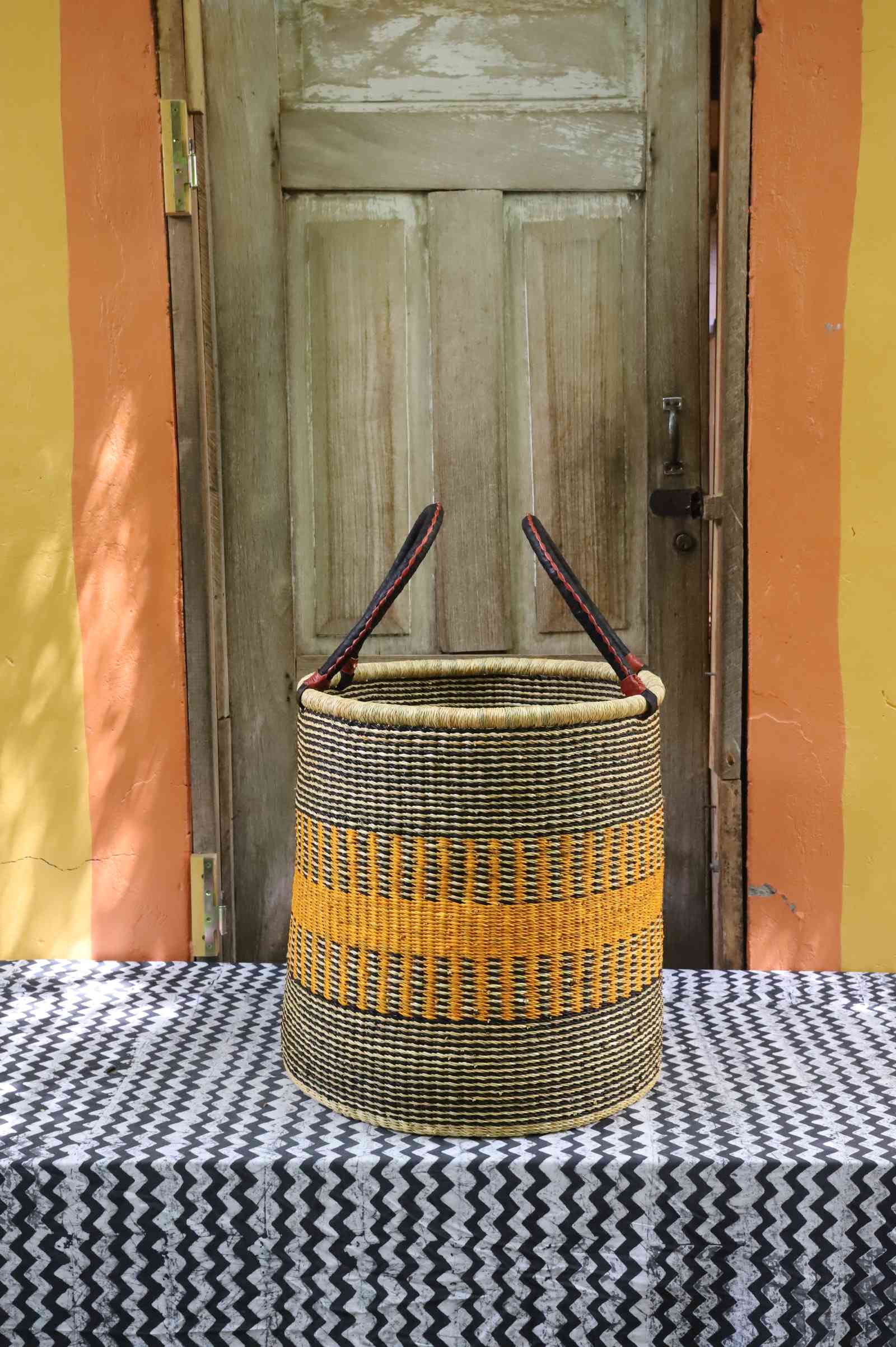 Tall open Bolga laundry basket in black and cream weave with a bold yellow stripe band and leather-topped handles.