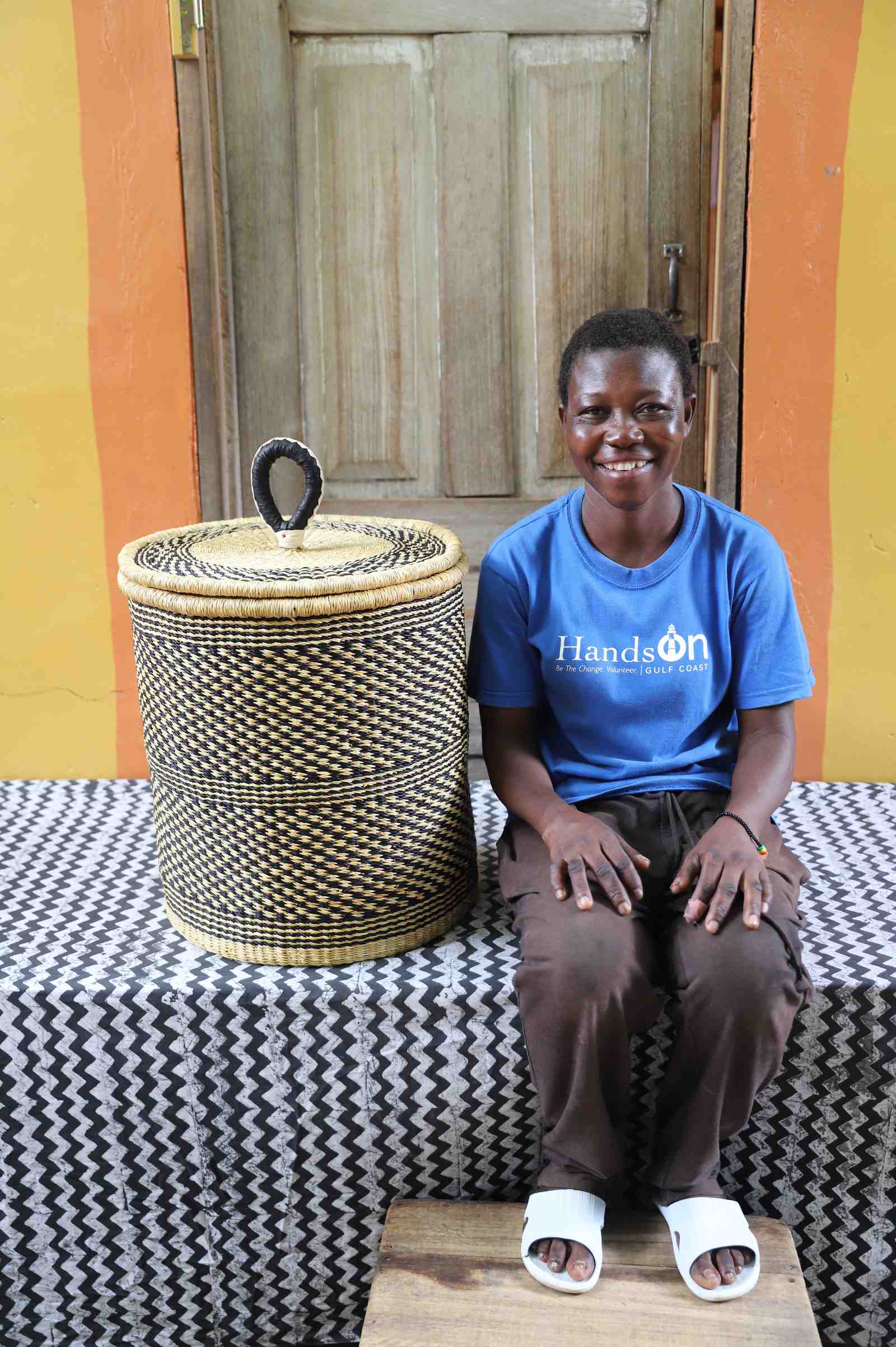 Coiled-rim lidded laundry basket in a black and tan speckled weave, shown beside a seated person for scale.
