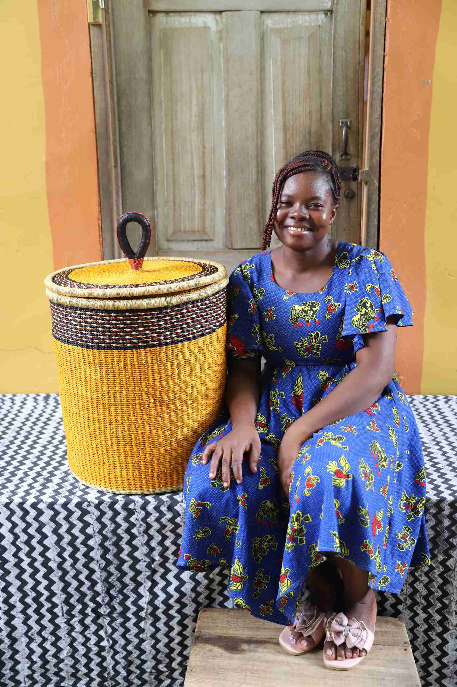 Coiled-rim lidded laundry basket in solid mustard weave, shown beside a seated person for scale.