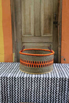 Handwoven Bolga storage basket in natural straw with an orange and black striped band and twin handles.