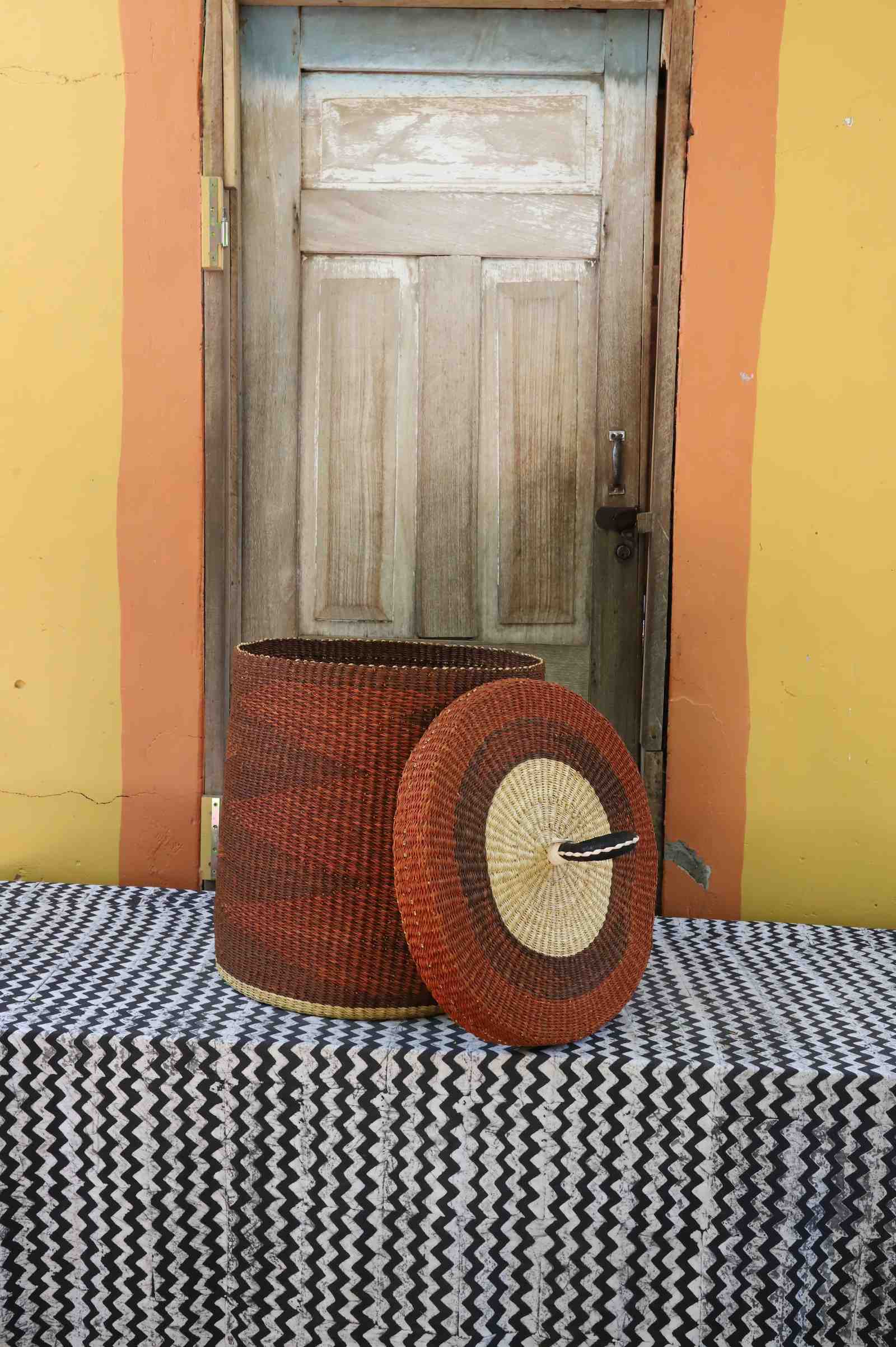 Handwoven Bolga laundry basket with lid removed, rust-red body with natural rim and matching lid.