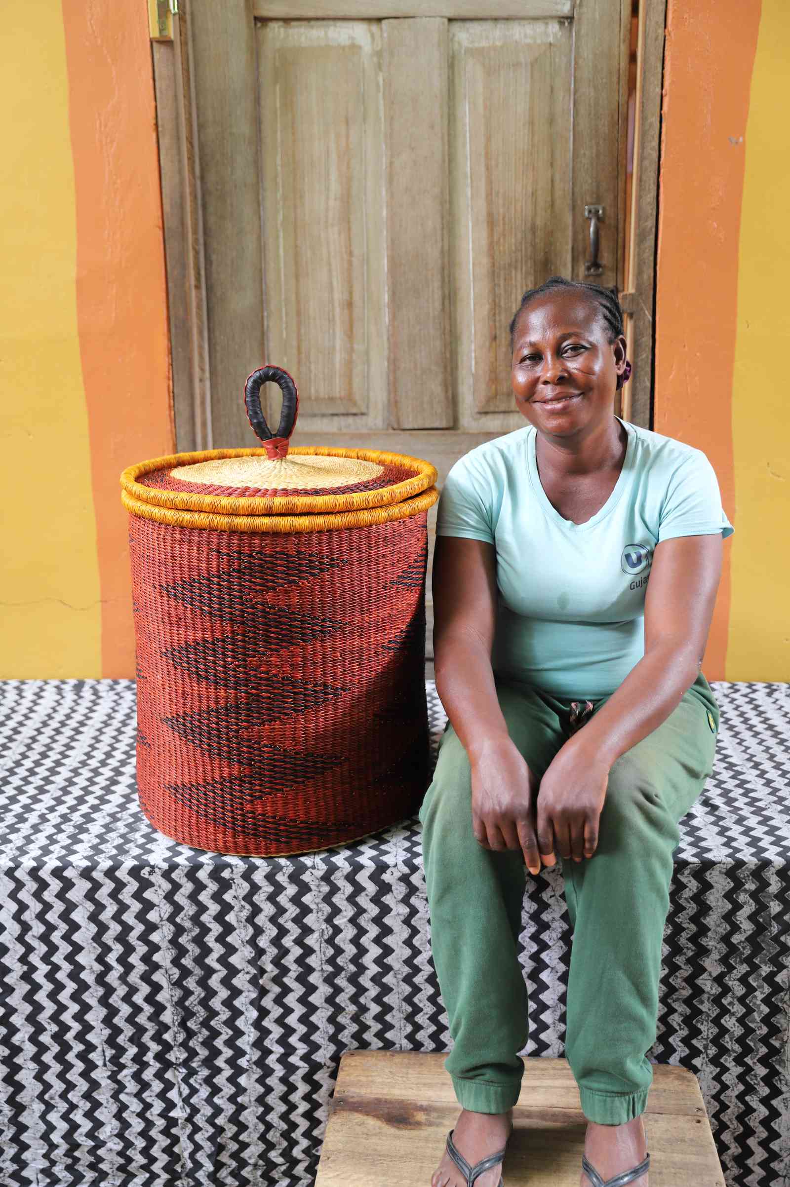 Coiled-rim lidded laundry basket in rust weave with a black diamond pattern, shown beside a seated person for scale.