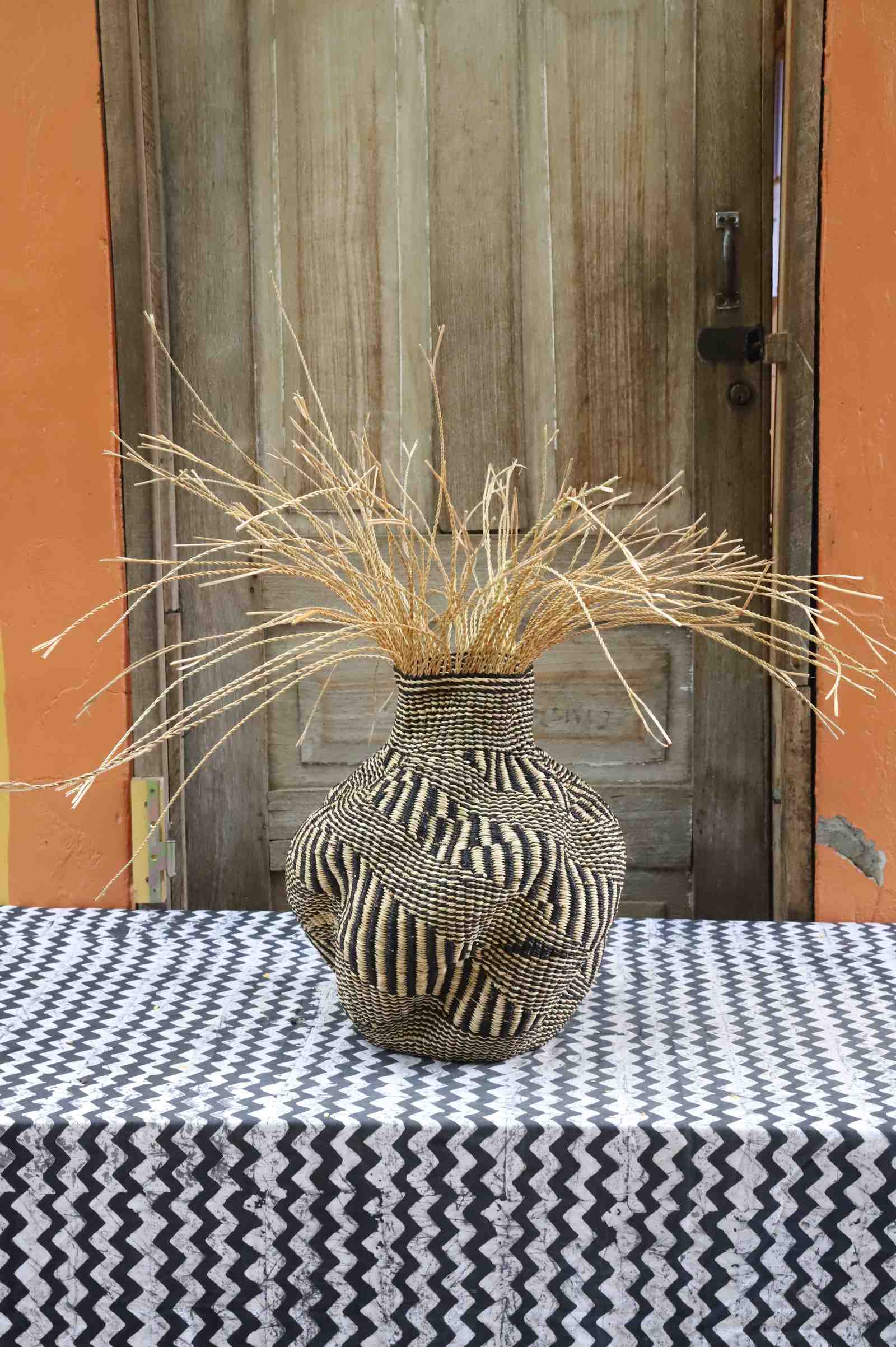 Handwoven Bolga Baobab-inspired basket in natural straw with a dark vertical trunk motif and natural grass sprigs.