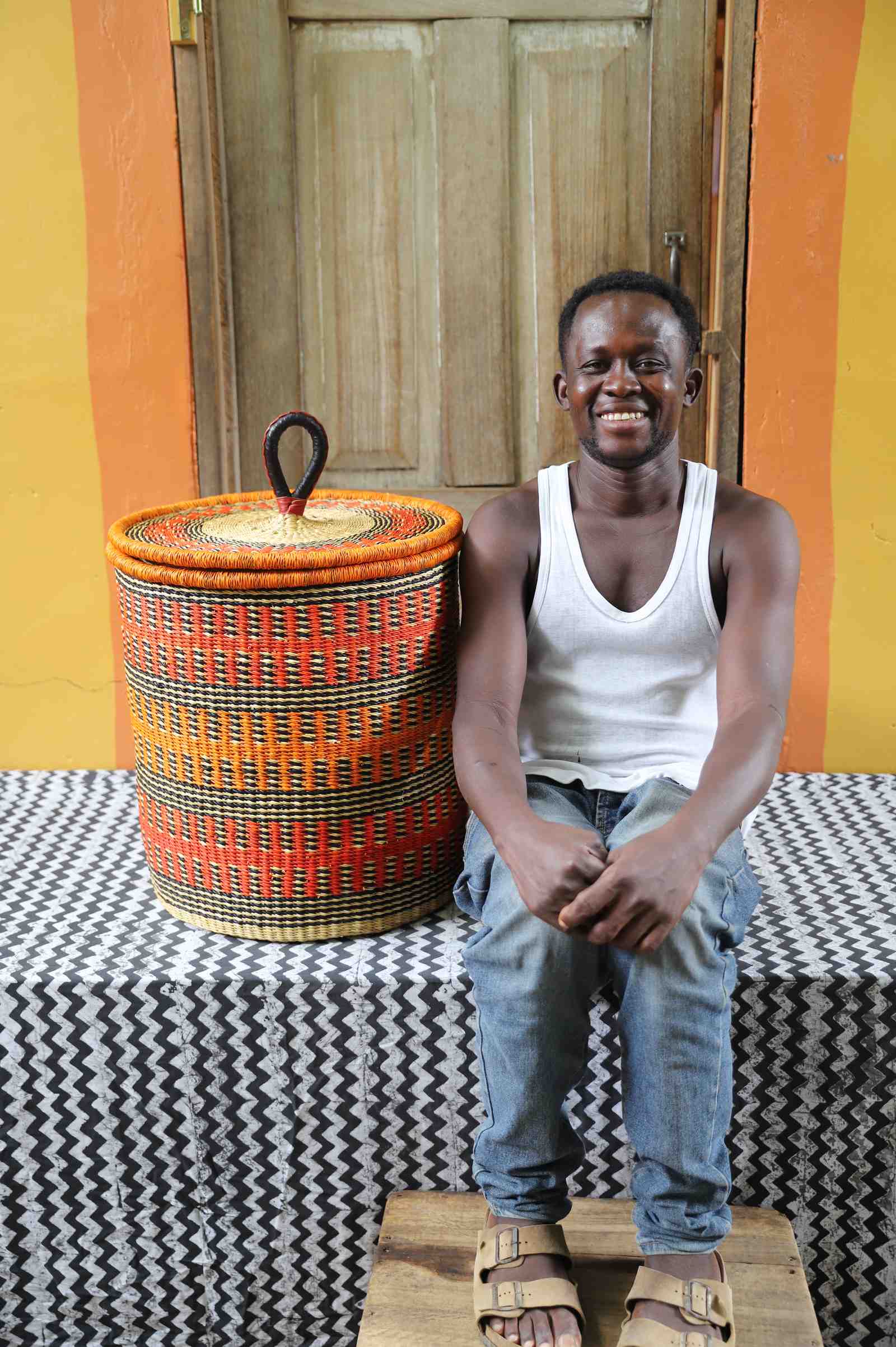 Coiled-rim lidded laundry basket in orange and black fine-striped weave, shown beside a seated person for scale.