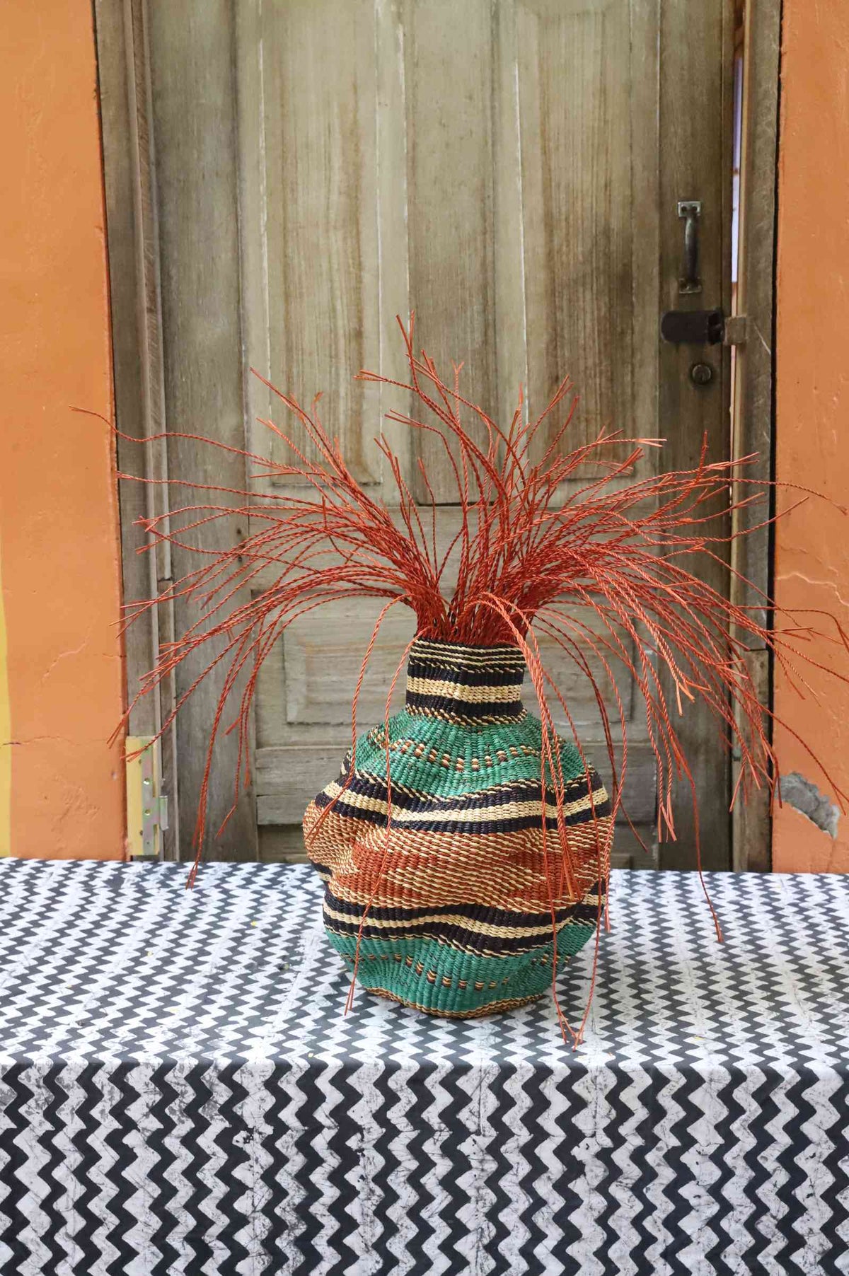 Handwoven Bolga Baobab-inspired basket in natural straw with orange and teal wave bands and rust-orange grass sprigs.