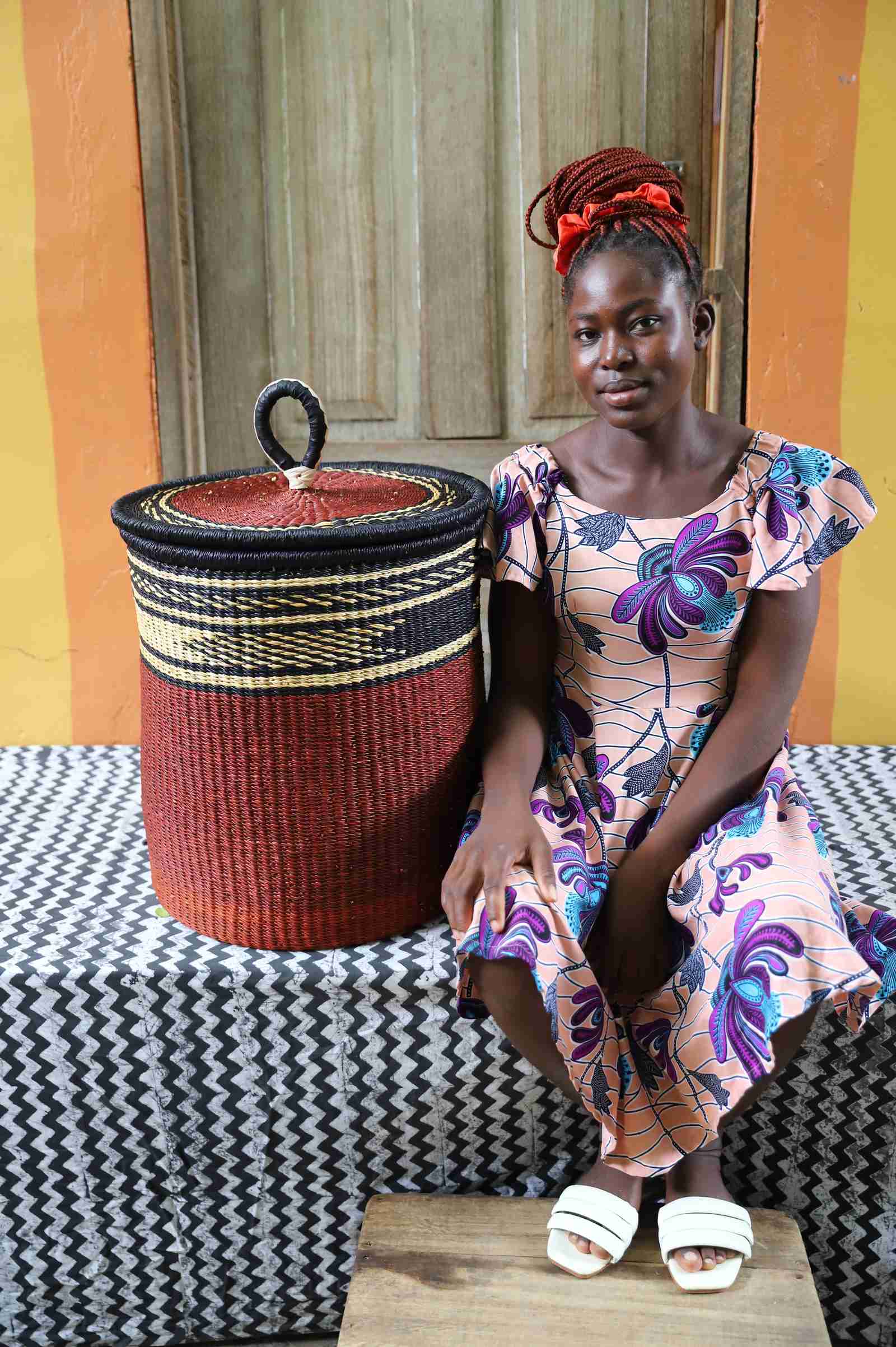 Coiled-rim lidded laundry basket in solid rust weave, shown beside a seated person for scale.