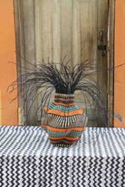 Handwoven Bolga Baobab-inspired basket in natural straw with wide orange and teal bands and dark grass sprigs.