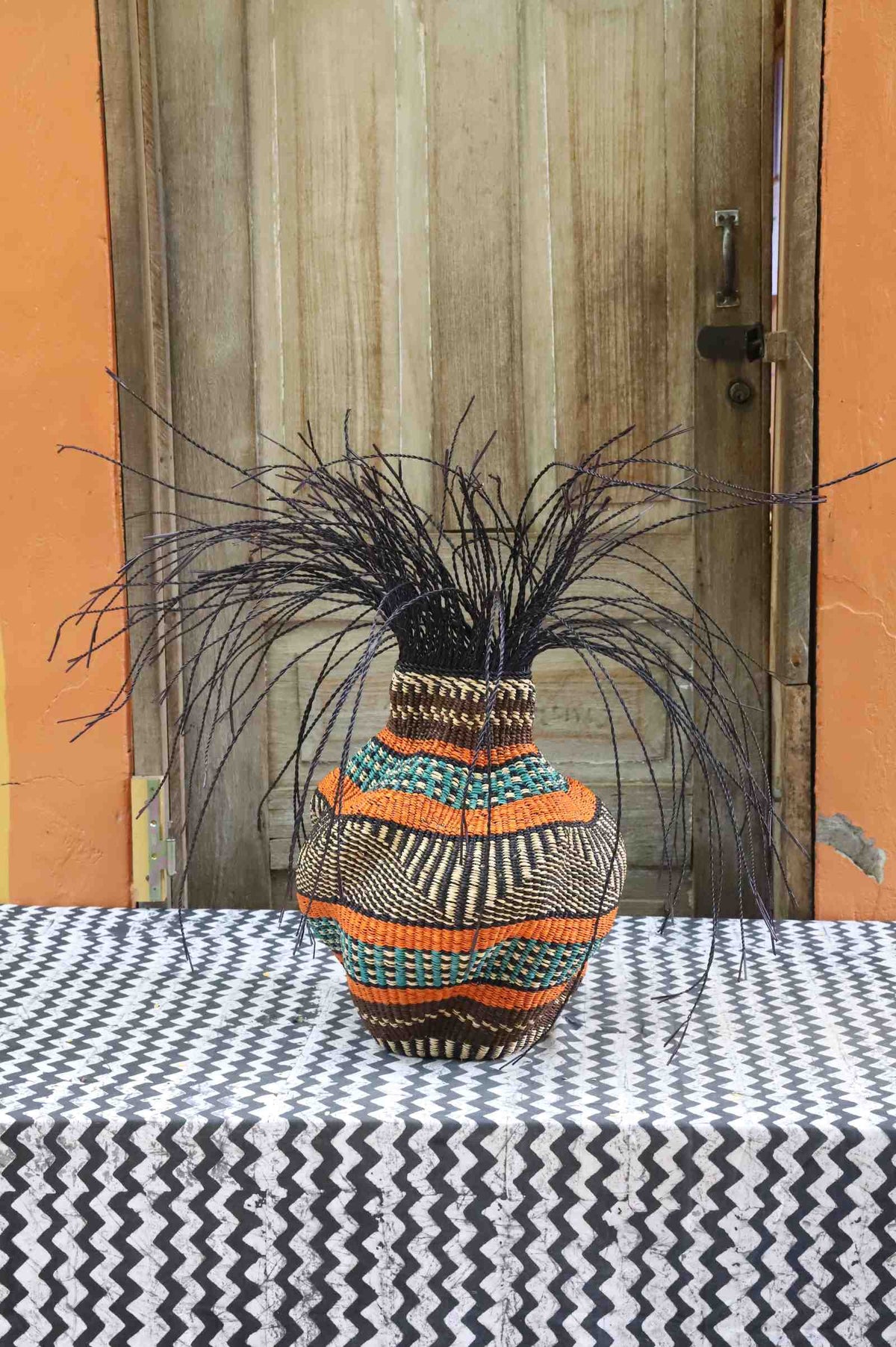 Handwoven Bolga Baobab-inspired basket in natural straw with wide orange and teal bands and dark grass sprigs.