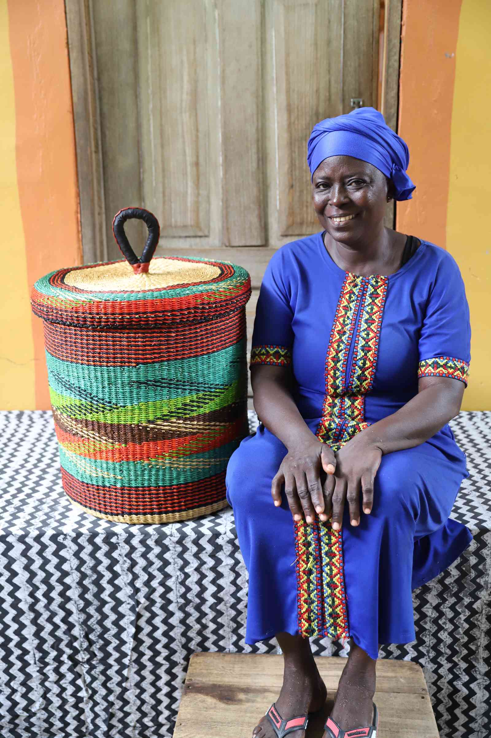 Lidded laundry basket in rust and teal wave-patterned weave, shown beside a seated person for scale.
