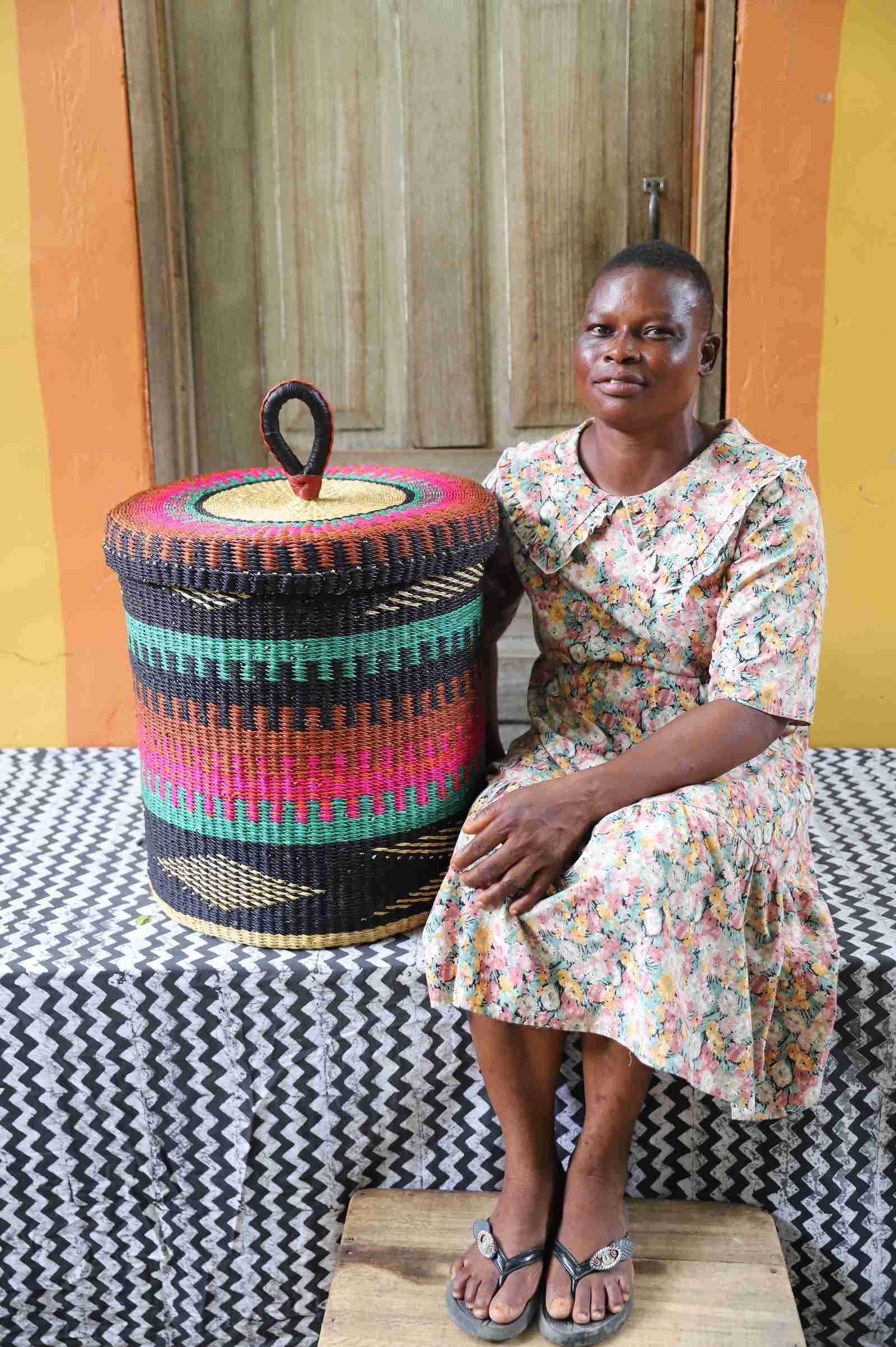 Lidded laundry basket in teal, pink and rust banded weave, shown beside a seated person for scale.