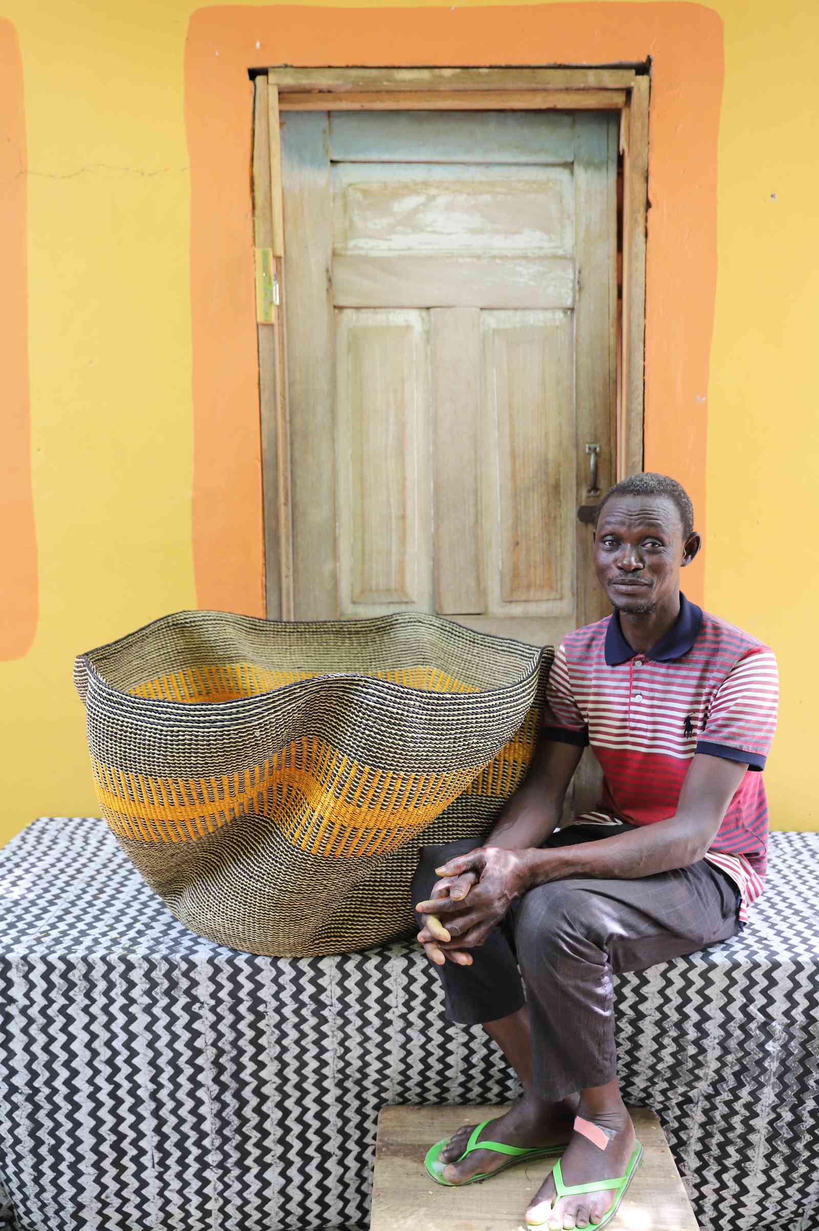 Large wave-form Bolga basket in grey and natural weave with an orange stripe, shown beside a seated man for scale.
