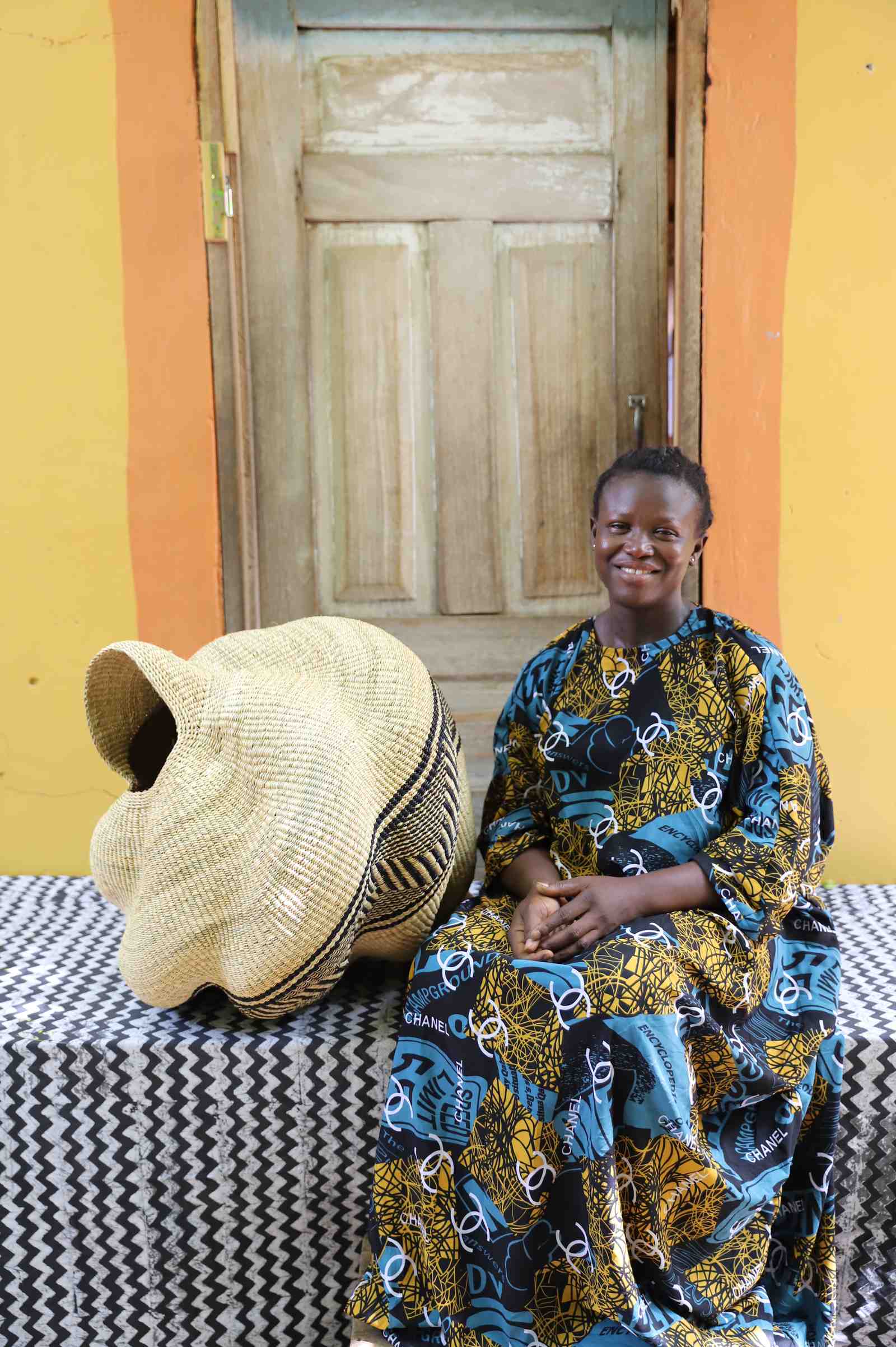 Natural straw Bolga basket in a spiral conch shape with a black geometric band, shown beside a seated woman for scale.