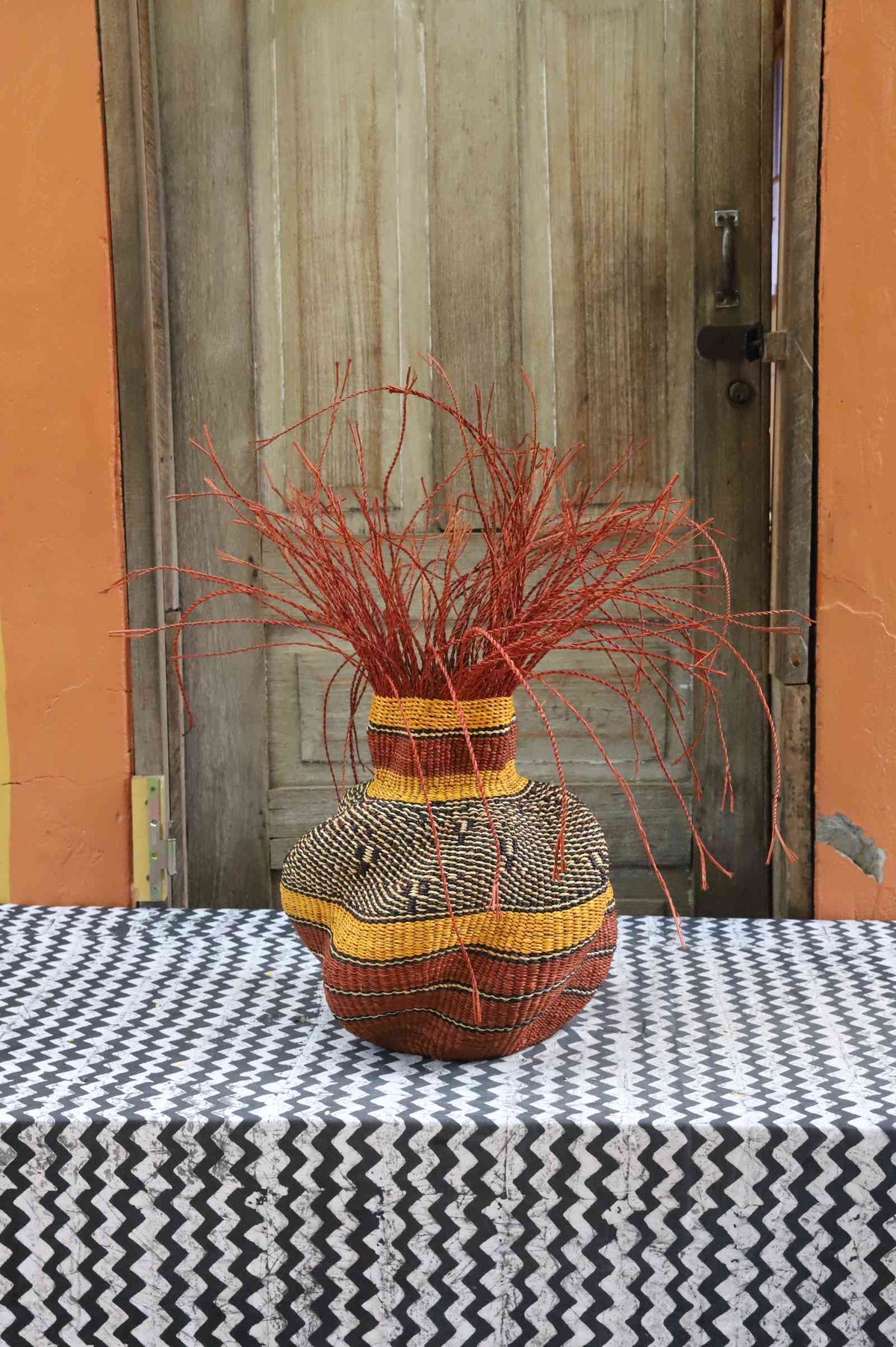 Handwoven Bolga Baobab-inspired basket in natural straw with orange banded shoulder and orange grass sprigs on top.