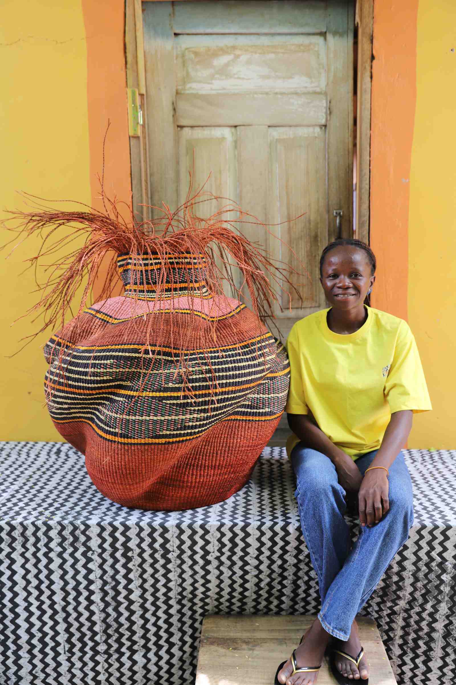 Large round Bolga basket in rust red with a striped band and loose straw fronds, shown beside a seated woman for scale.