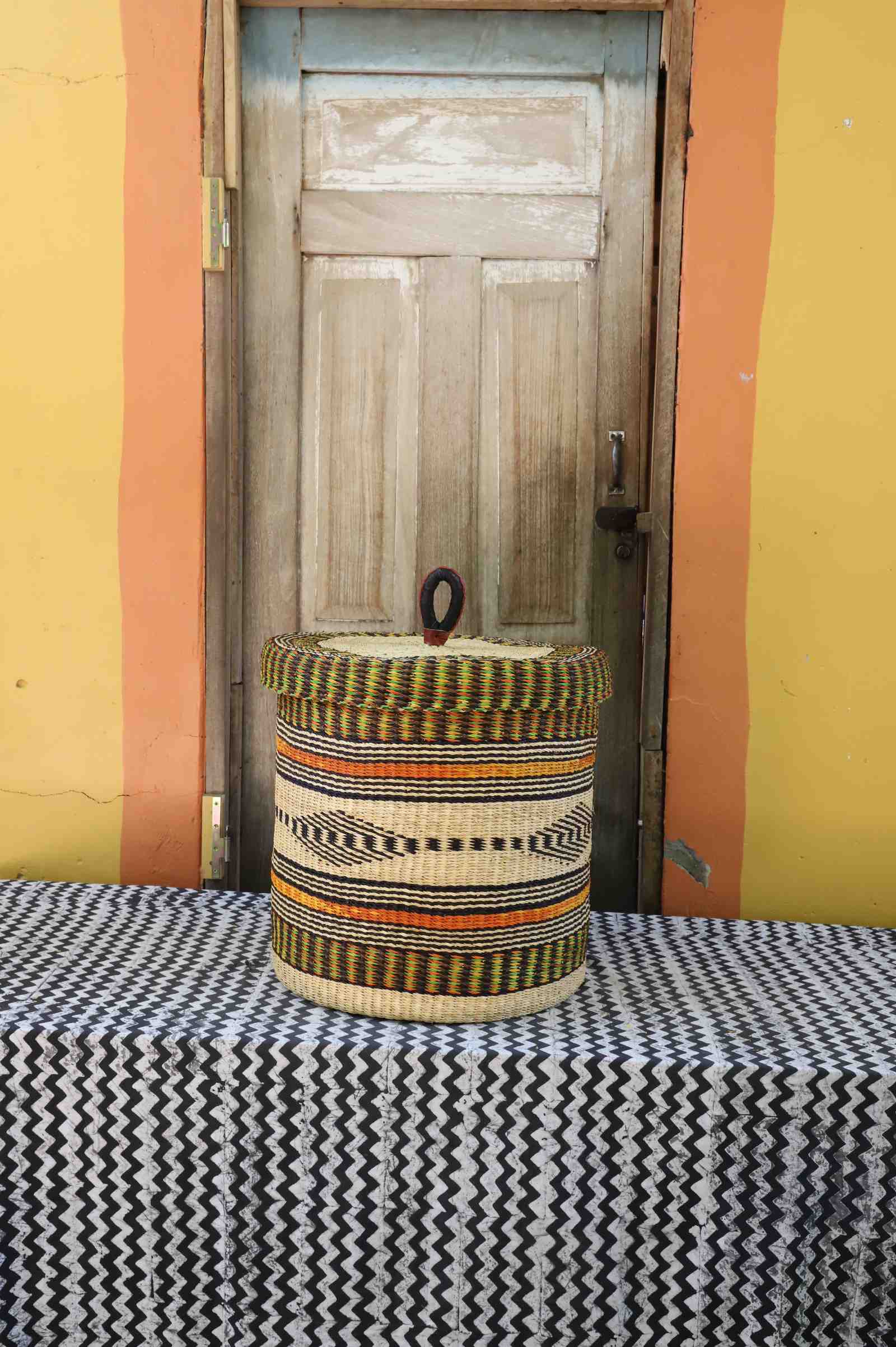 Handwoven Bolga laundry basket with lid in natural straw with black, red, orange and yellow banded stripes.