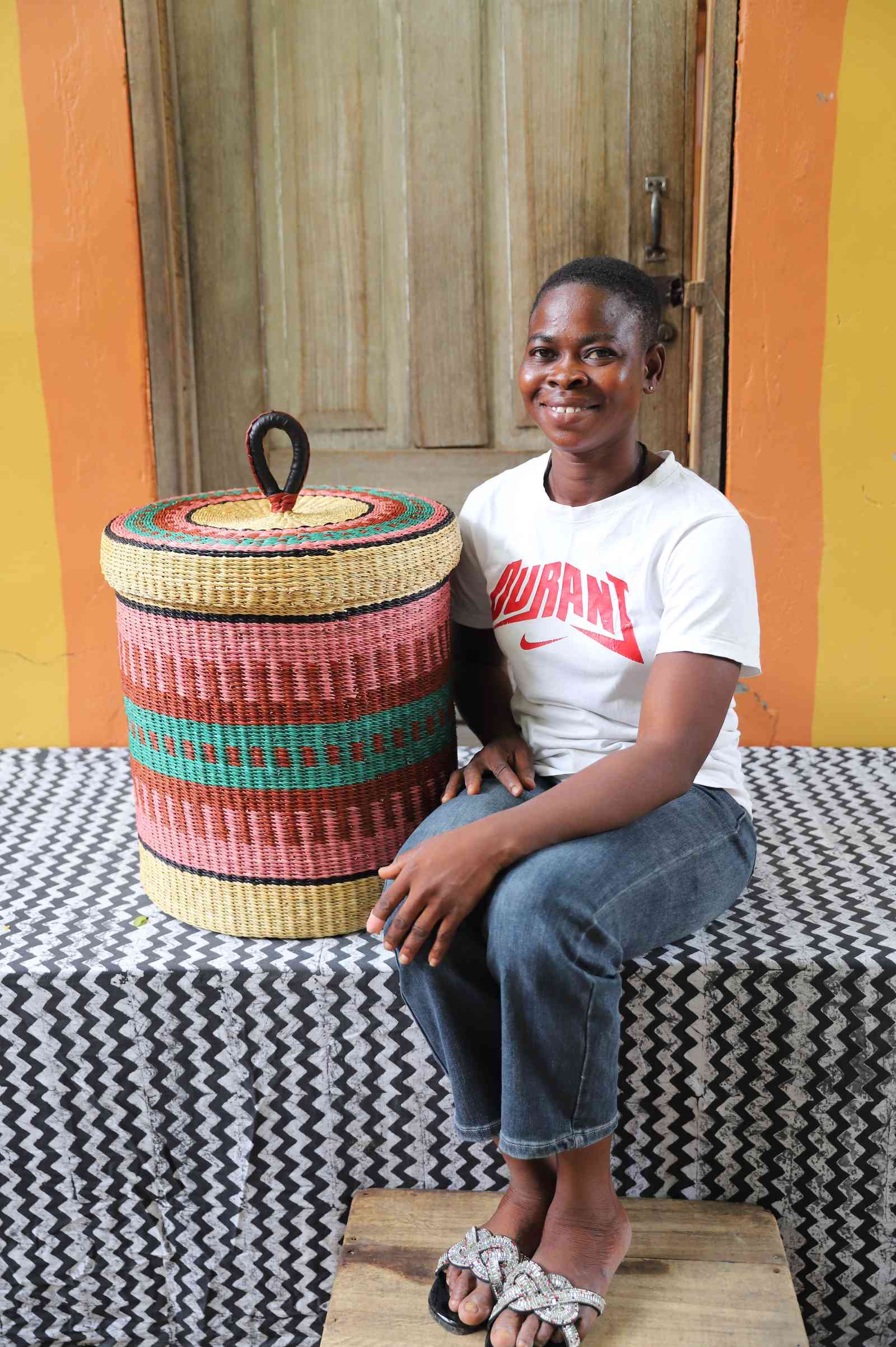 Lidded laundry basket in natural straw with pink and teal striped banding, shown beside a seated person for scale.