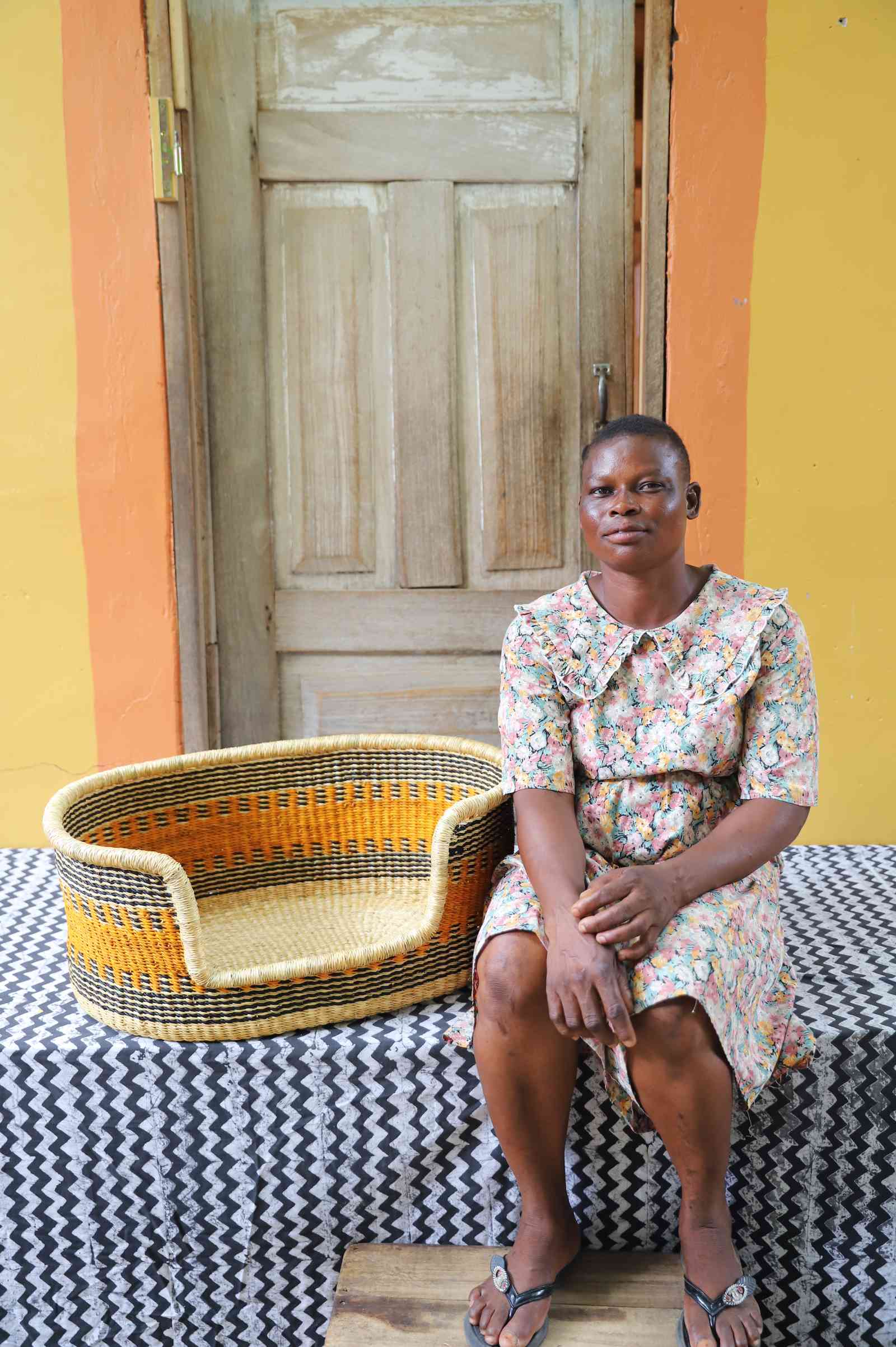 Woven dog bed in natural straw with an orange and dark checkered pattern, shown beside a seated person for scale.