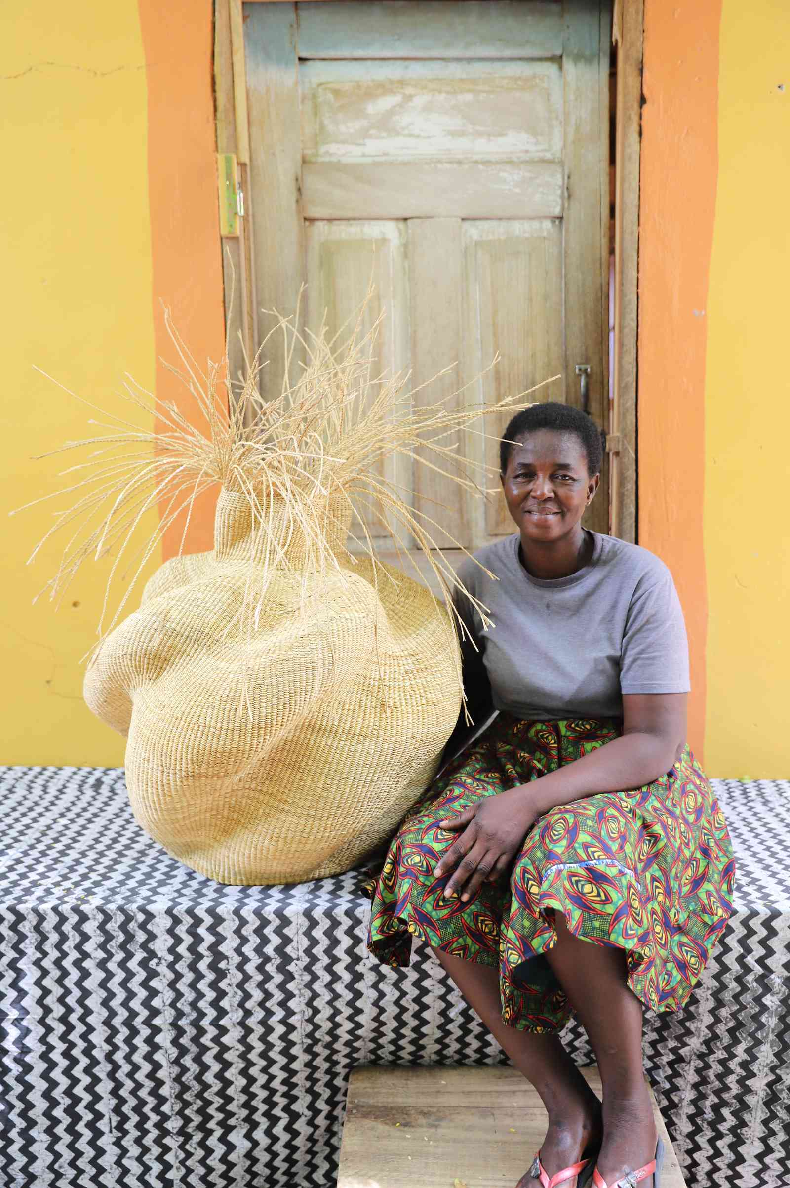 Round Bolga basket in natural straw with loose straw fronds at the neck, shown beside a seated woman for scale.