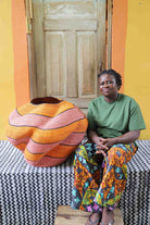 Large nest-shaped Bolga art basket in pink and rust wavy braided weave, shown beside a seated person for scale.