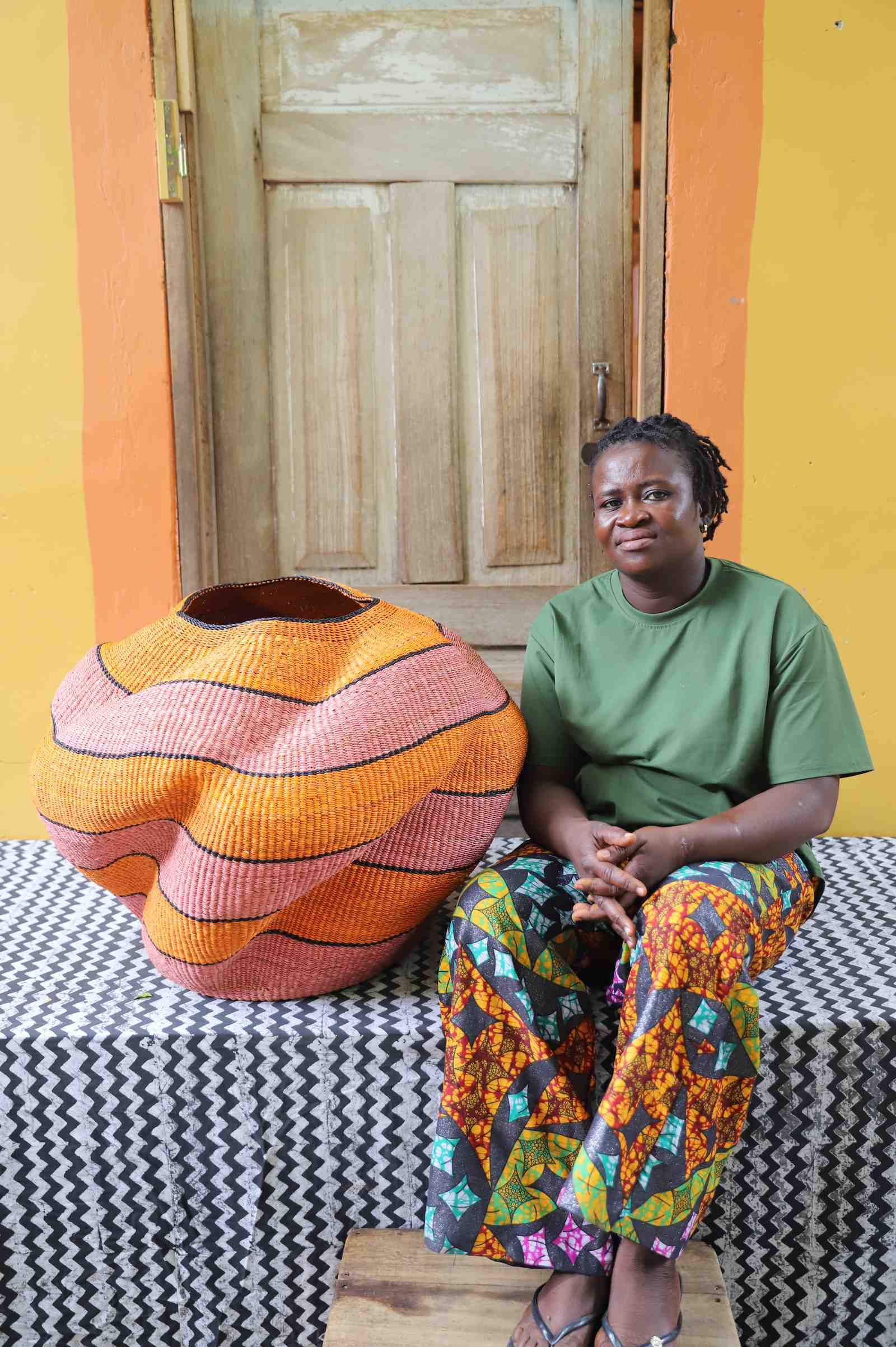 Large nest-shaped Bolga art basket in pink and rust wavy braided weave, shown beside a seated person for scale.