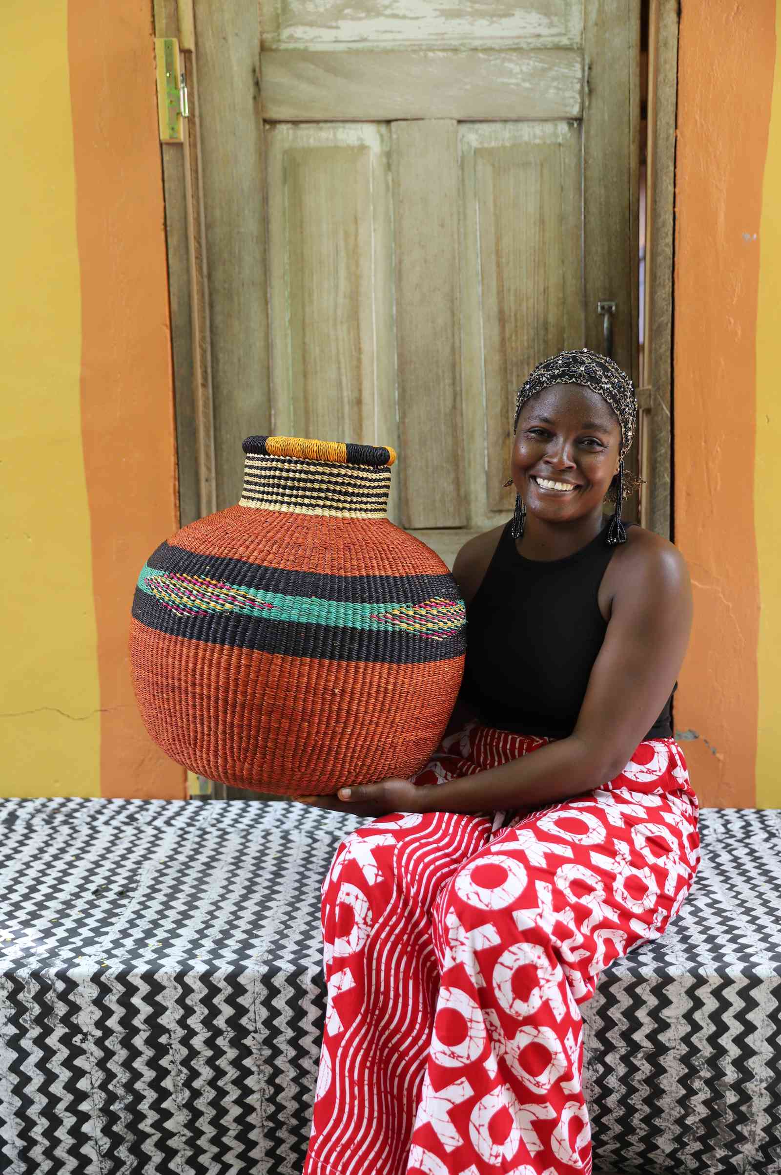 Round terracotta Bolga basket with a striped neck and green diamond band, held by a seated woman for scale.