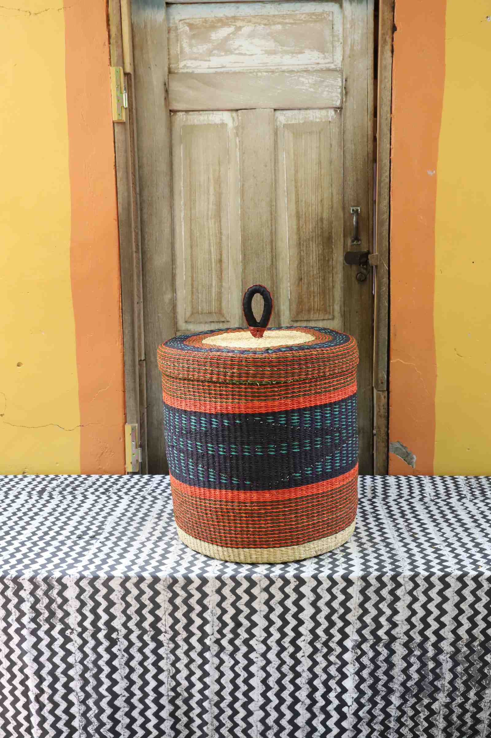Large handwoven Bolga laundry basket with lid in rust-orange with a dark central band and rust-red stripes.