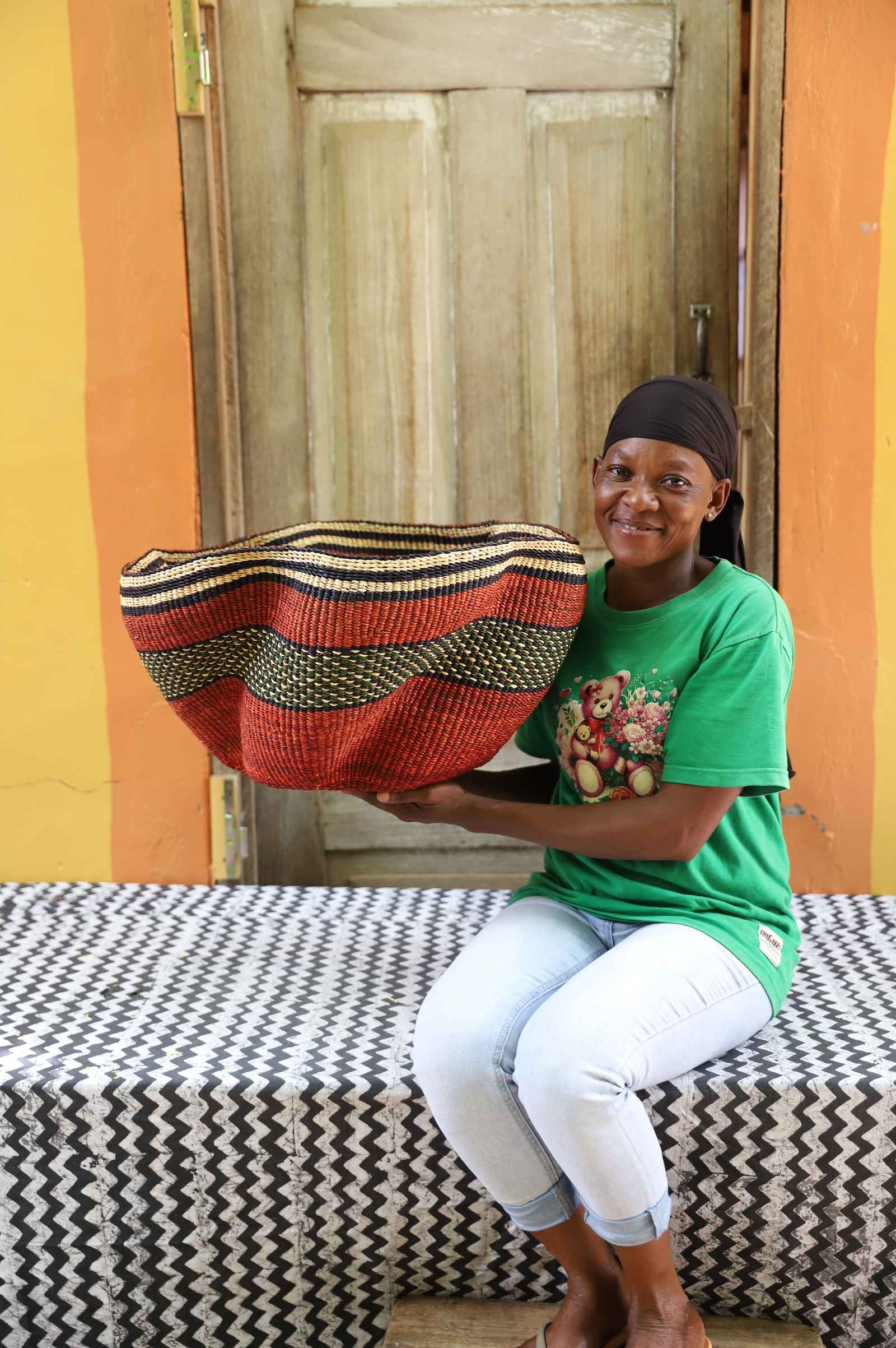 Wide rust red Bolga basket with a wavy rim and black-and-gold wave band, held by a seated woman for scale.