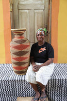 Tall vase-shaped Bolga art basket in natural straw with a brown and rust diamond pattern, shown beside a seated person for scale.