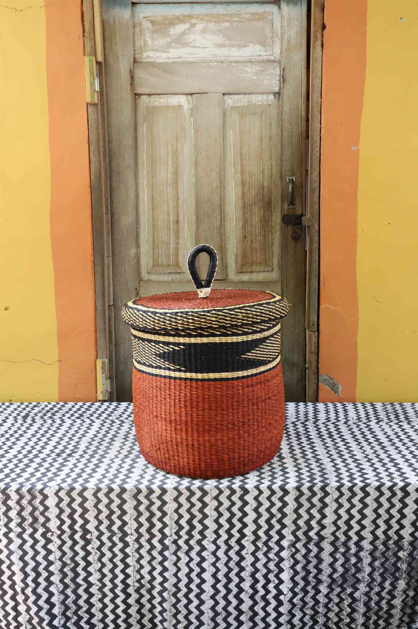 Medium handwoven Bolga laundry basket with lid, dark top with rust-orange banded base and knob handle.