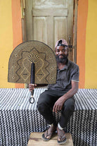 Naba Penga wall fan in dark brown straw with a tan star-shaped geometric weave, held by a seated person for scale.