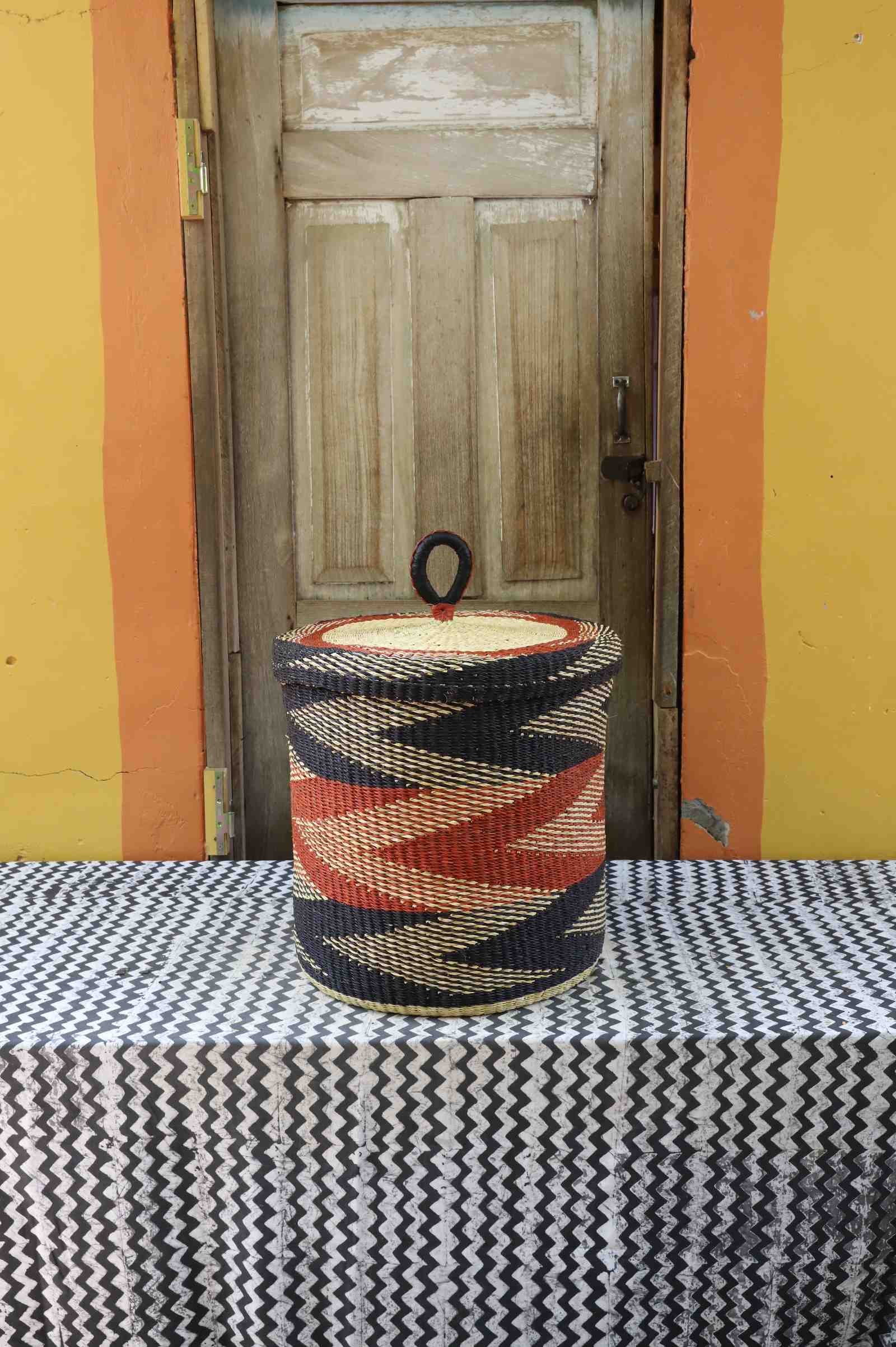 Medium handwoven Bolga laundry basket with lid in natural straw with rust-red and black banded stripes.