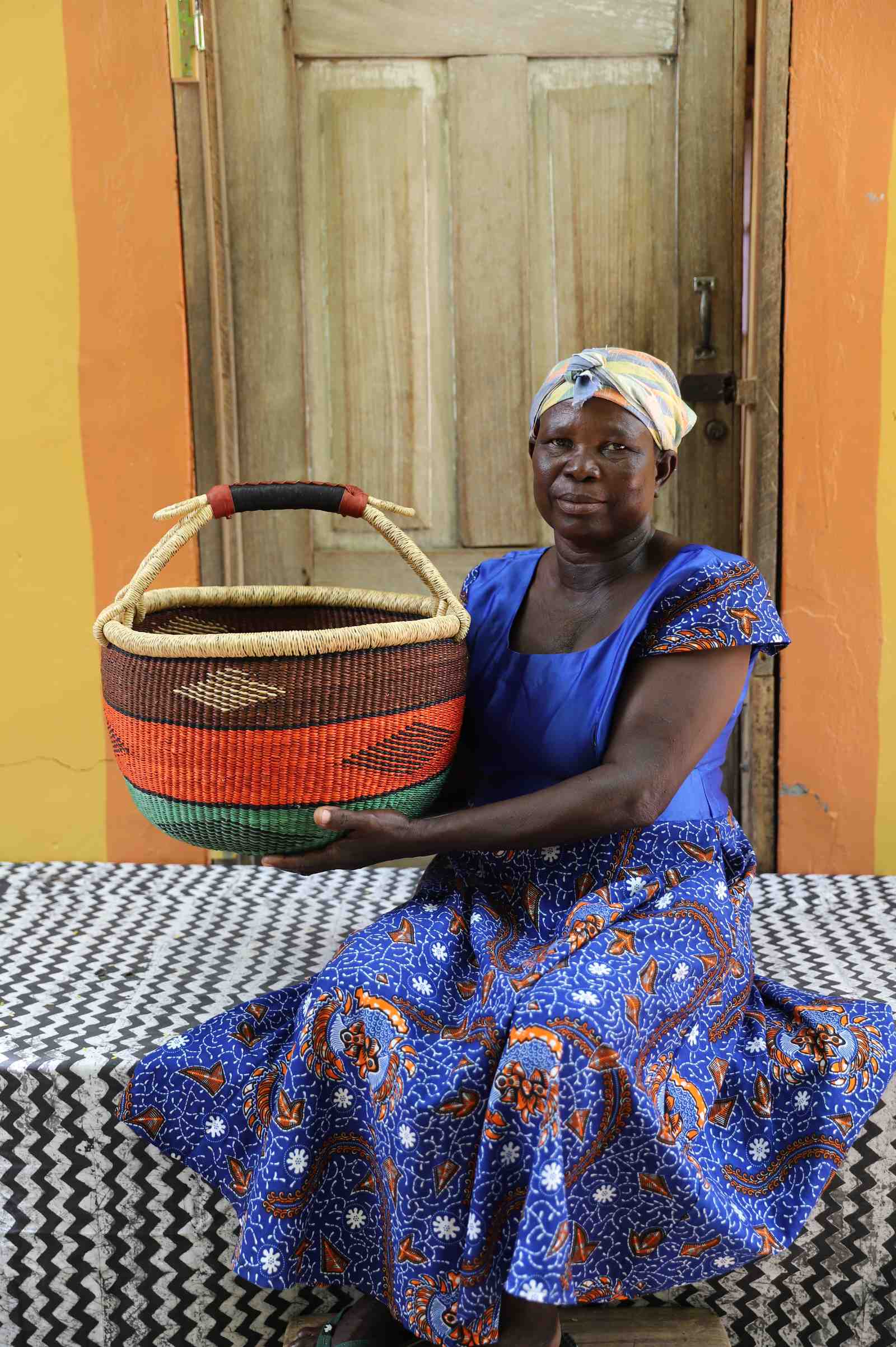 Round Bolga shopping basket striped in brown, orange and green with a leather-wrapped handle, held by a seated woman.