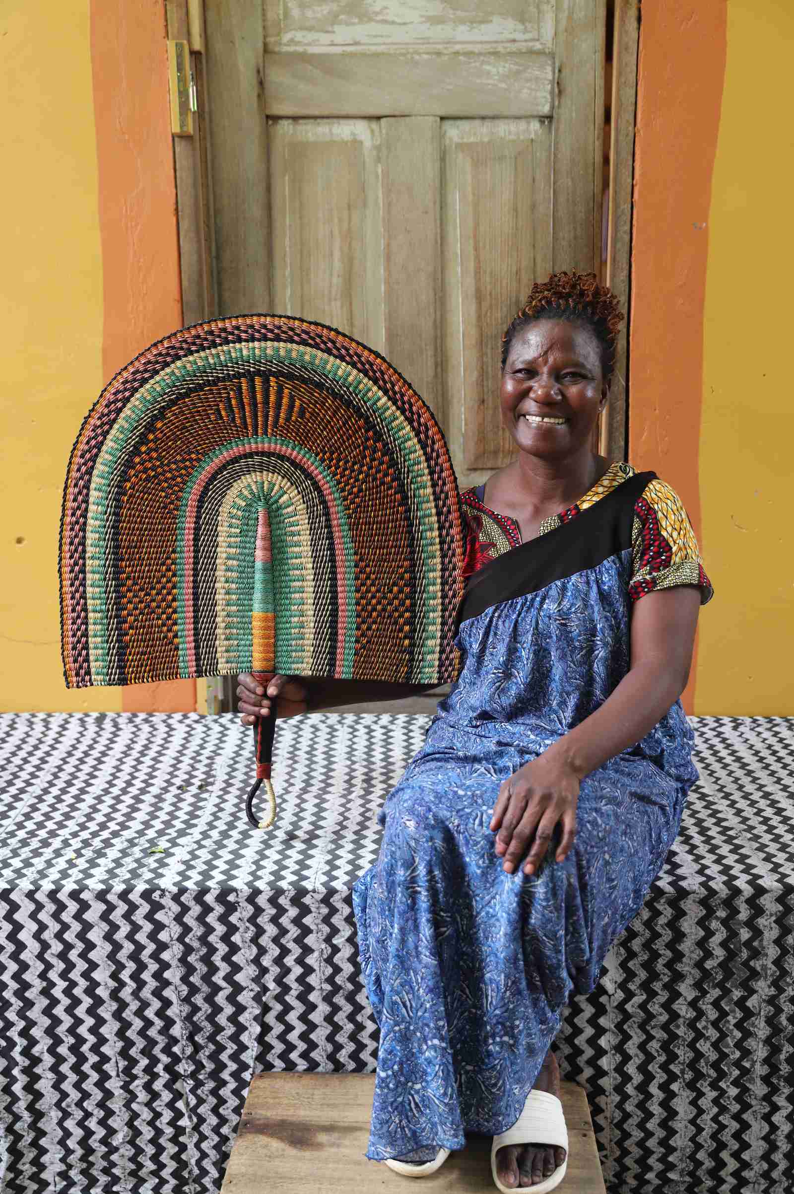 Naba Penga wall fan in natural straw with sage green and rust banded arches, held by a seated person for scale.