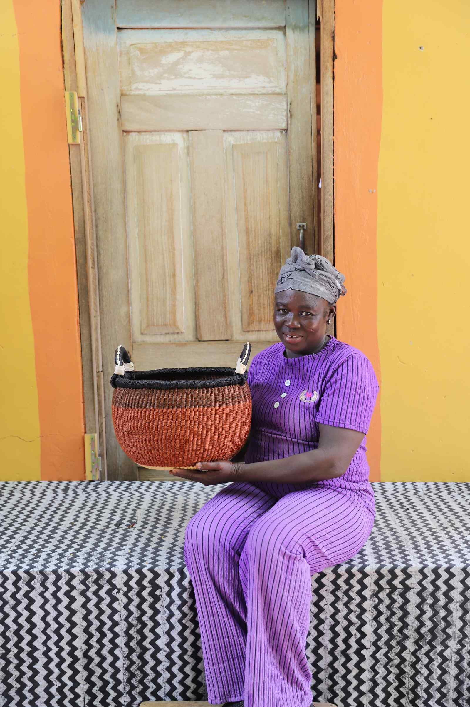 Handwoven Gbenka Bolga basket in rust with black rim, held by a woman seated against a yellow ochre wall in Bolgatanga.