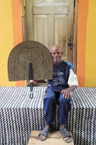 Naba Penga wall fan in natural straw with a dark geometric diamond weave, held by a seated person for scale.
